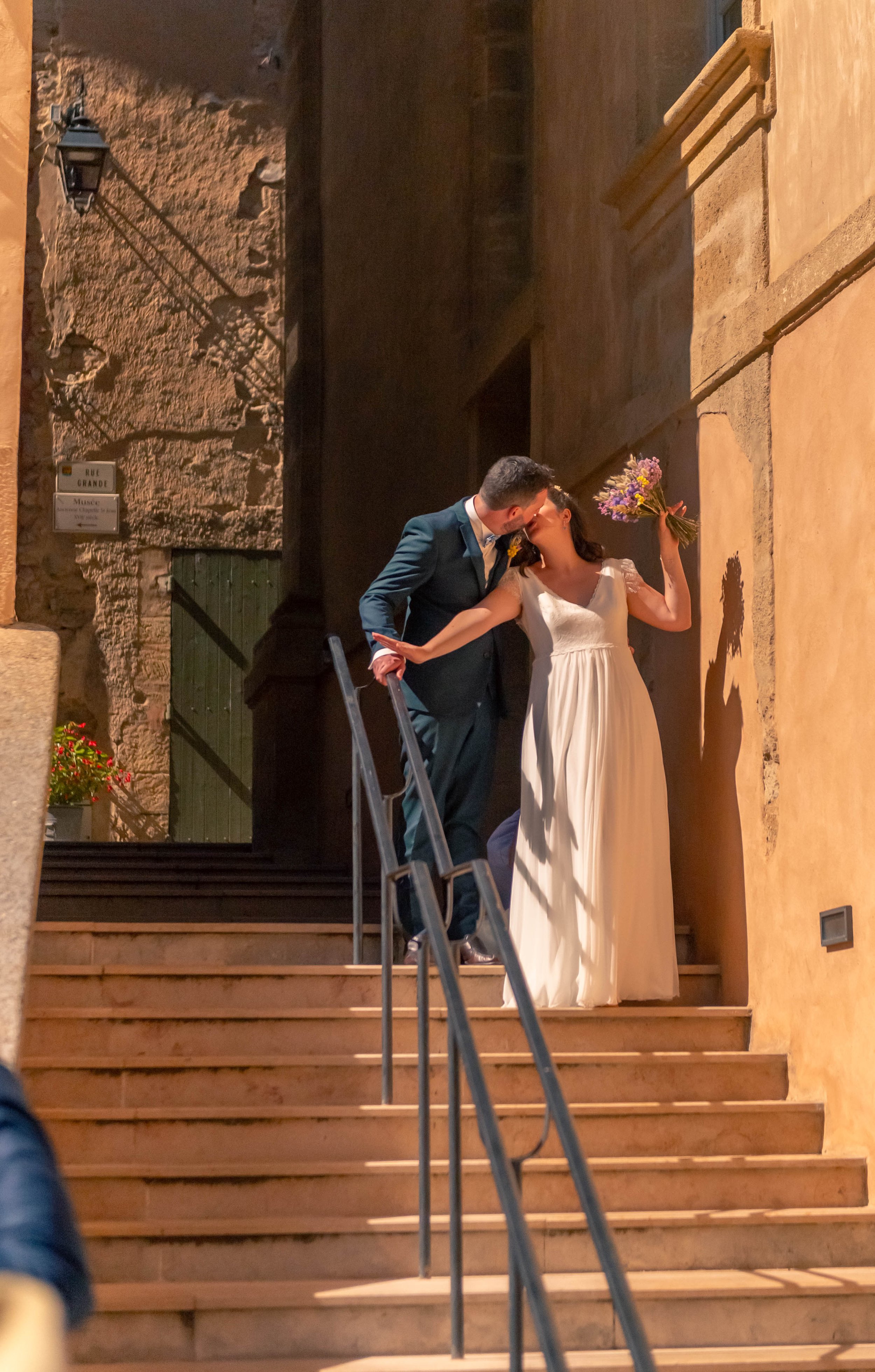 Un couple marié partage un baiser sur un escalier en pierre dans une rue pavée, la mariée tient un bouquet de fleurs, un décor romantique.