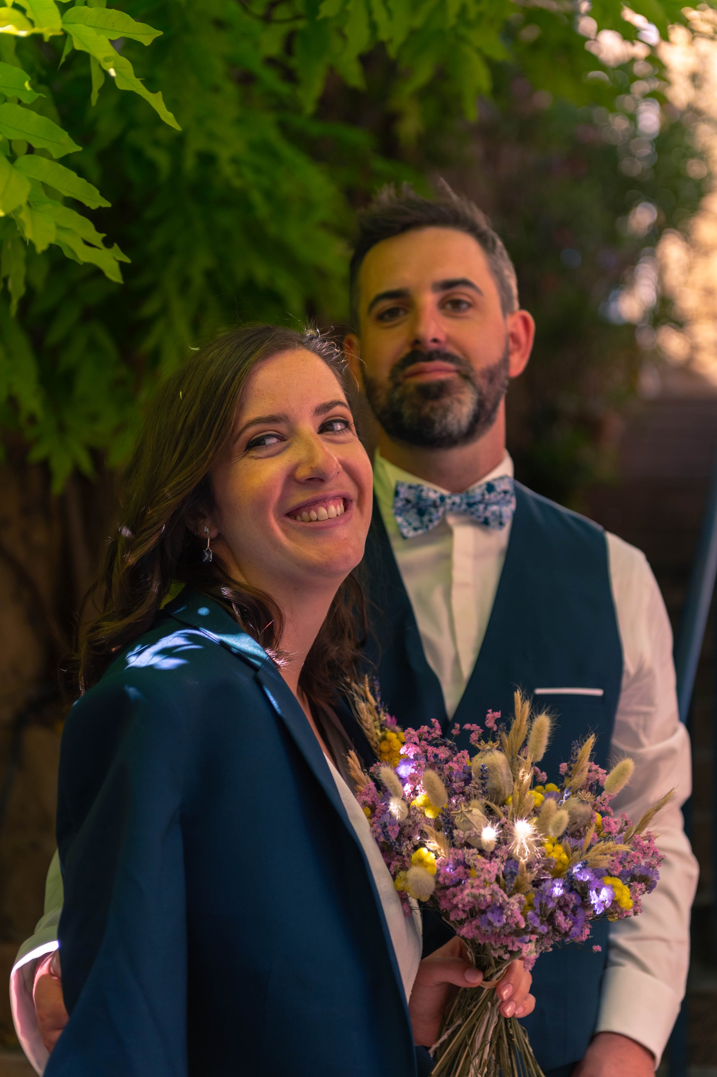 Un couple souriant lors d'une occasion spéciale, la femme tenant un bouquet de fleurs colorées, en extérieur avec un fond de feuillage.