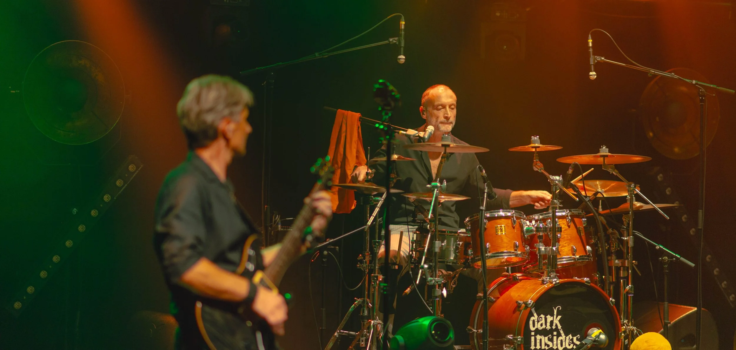 Deux musiciens jouent sur scène. Un guitariste à gauche, légèrement flou, et un batteur au centre, plus net, derrière sa batterie. La scène est éclairée de lumière chaude, avec un fond sombre.