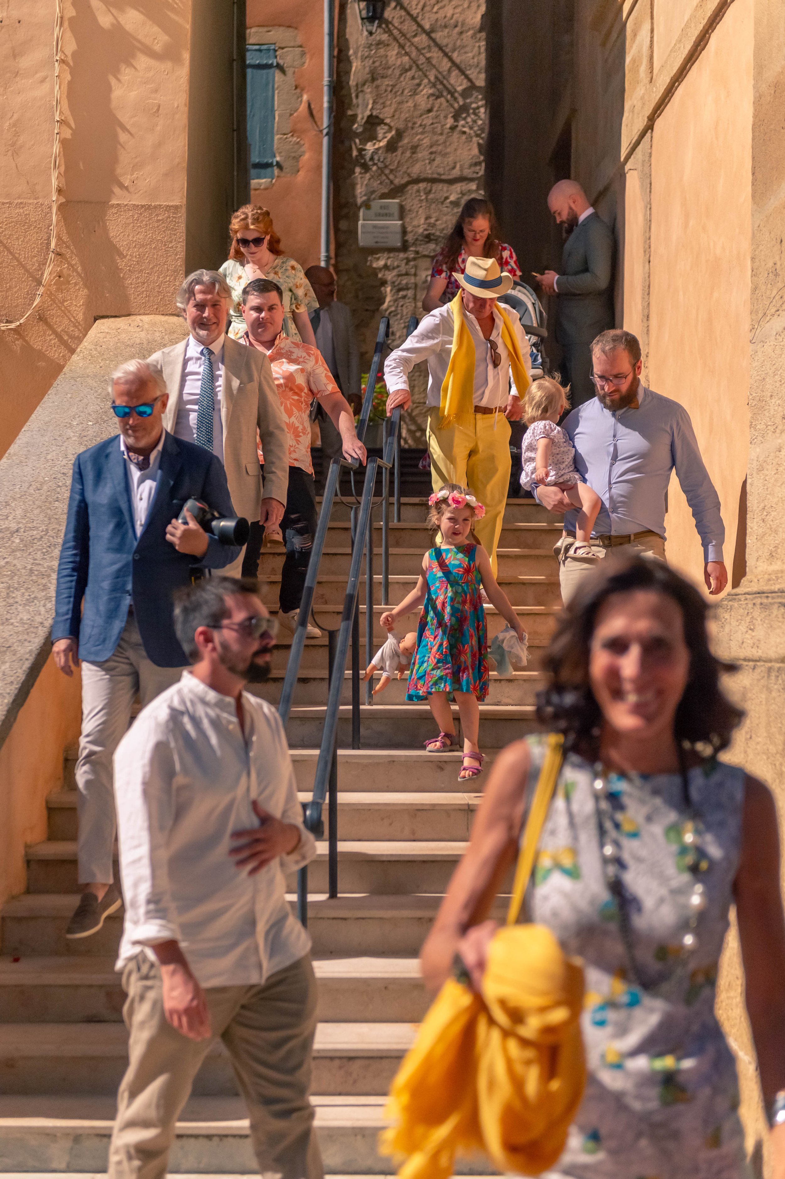 Groupe de personnes descendant un escalier en pierre sous un éclairage chaud, avec des murs en pierre et des bâtiments en couleur terre cuite en arrière-plan, lors d'une soirée ou d'un événement.