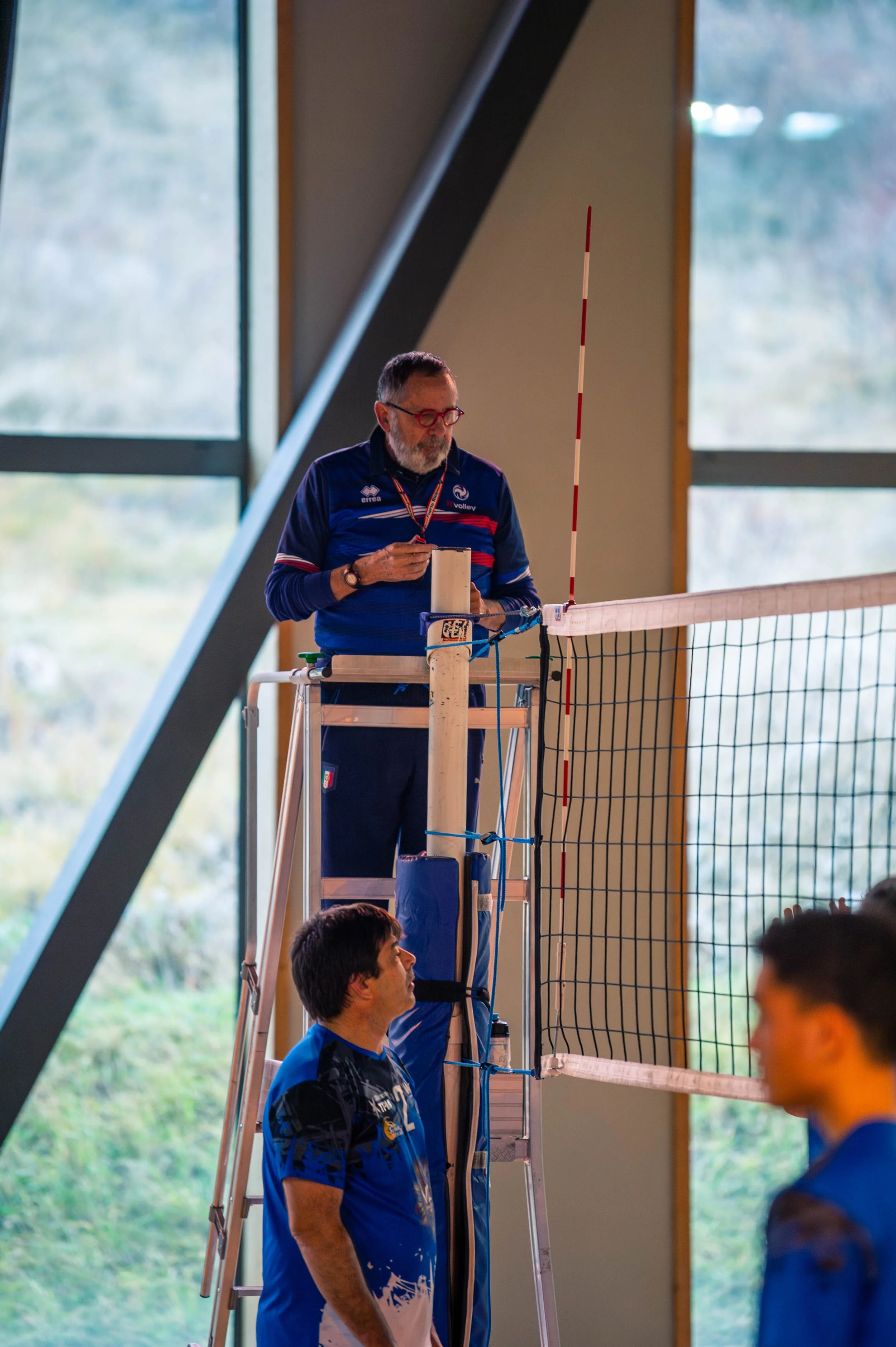 Un arbitre de volleyball observe une équipe lors d'un match à l'intérieur d'un gymnase, avec deux joueurs en bleu devant le filet.