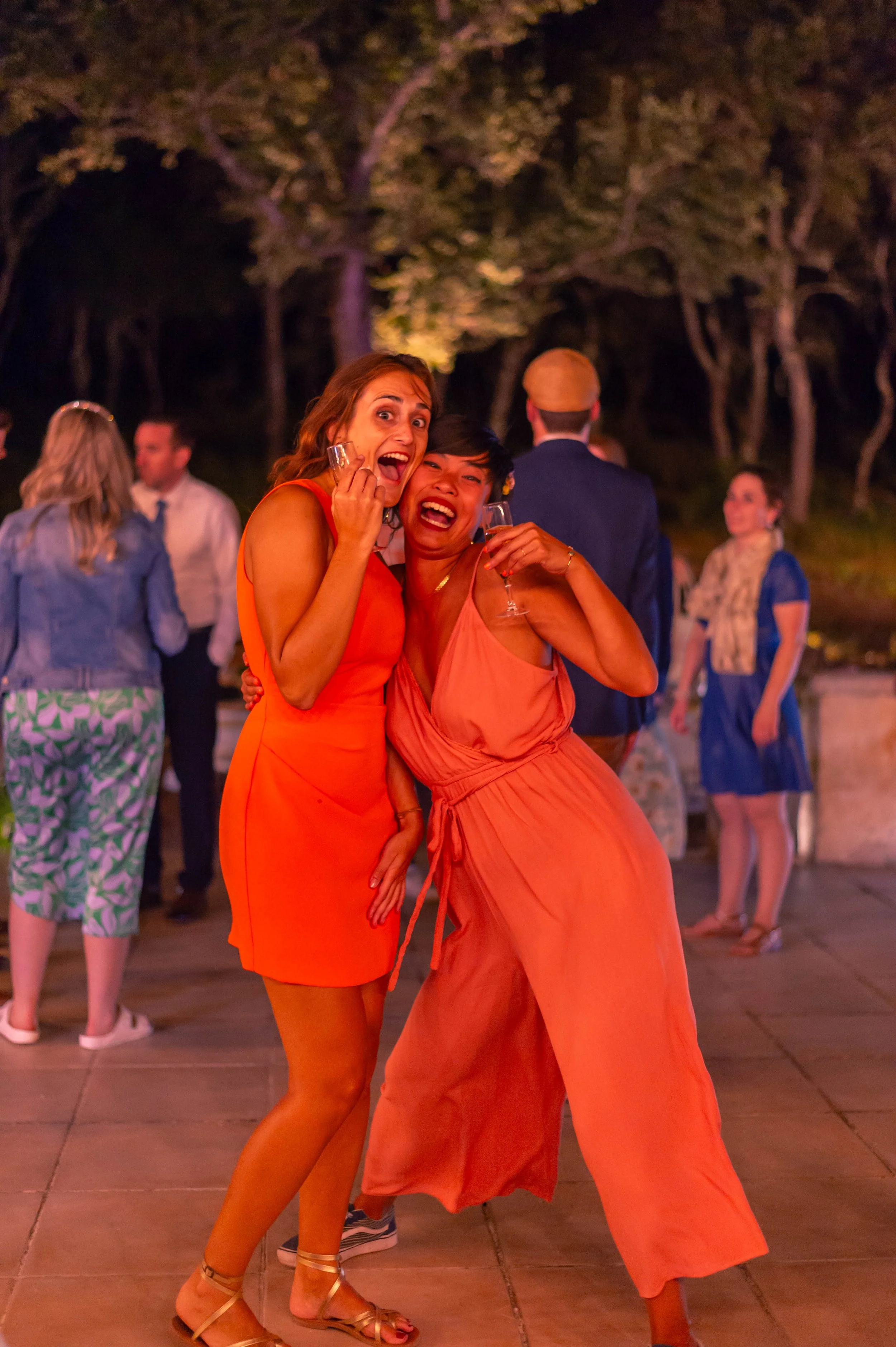 Deux femmes souriantes lors d'une fête en extérieur, tenant des verres de champagne, vêtues de robes orange, entourées d'autres personnes en arrière-plan.