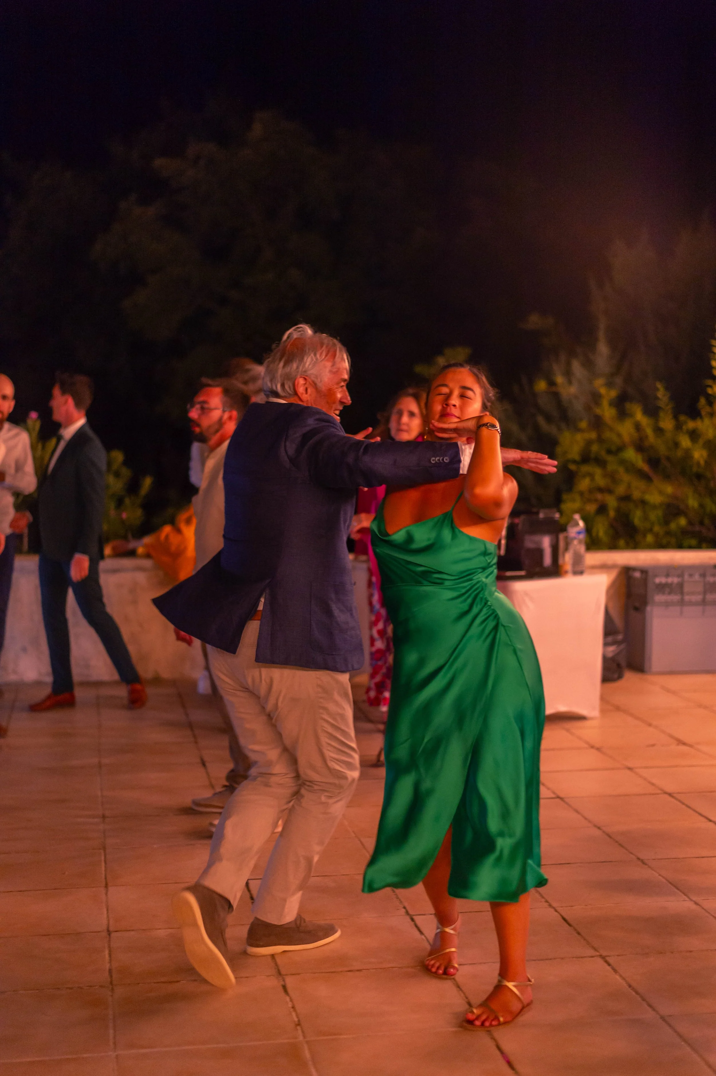 Un couple danse lors d'une fête en plein air la nuit, avec plusieurs personnes en arrière-plan.