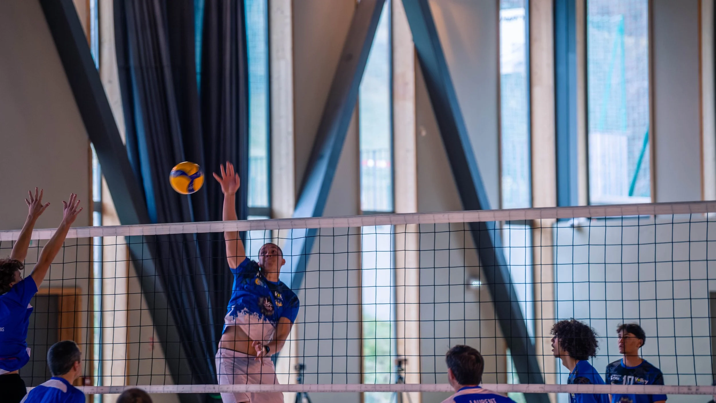 Une partie de volleyball en intérieur, un joueur en bleu saute au filet pour frapper la balle tandis que ses coéquipiers et adversaires se préparent en arrière.