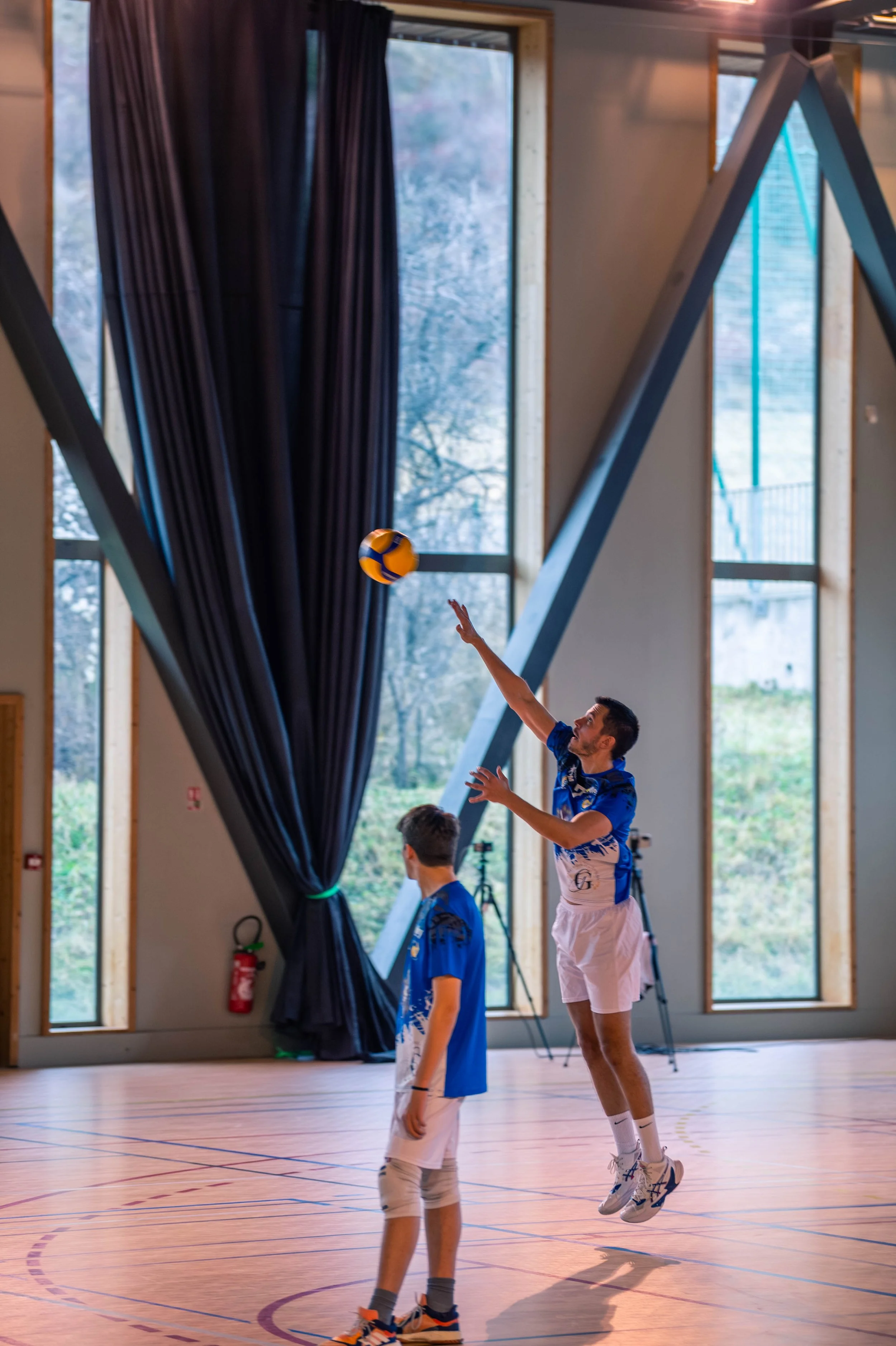 deux jeunes hommes jouent au volleyball en intérieur, un saut attemptant de frapper la balle, portant des maillots bleus, dans une salle avec grandes fenêtres et rideaux noirs.