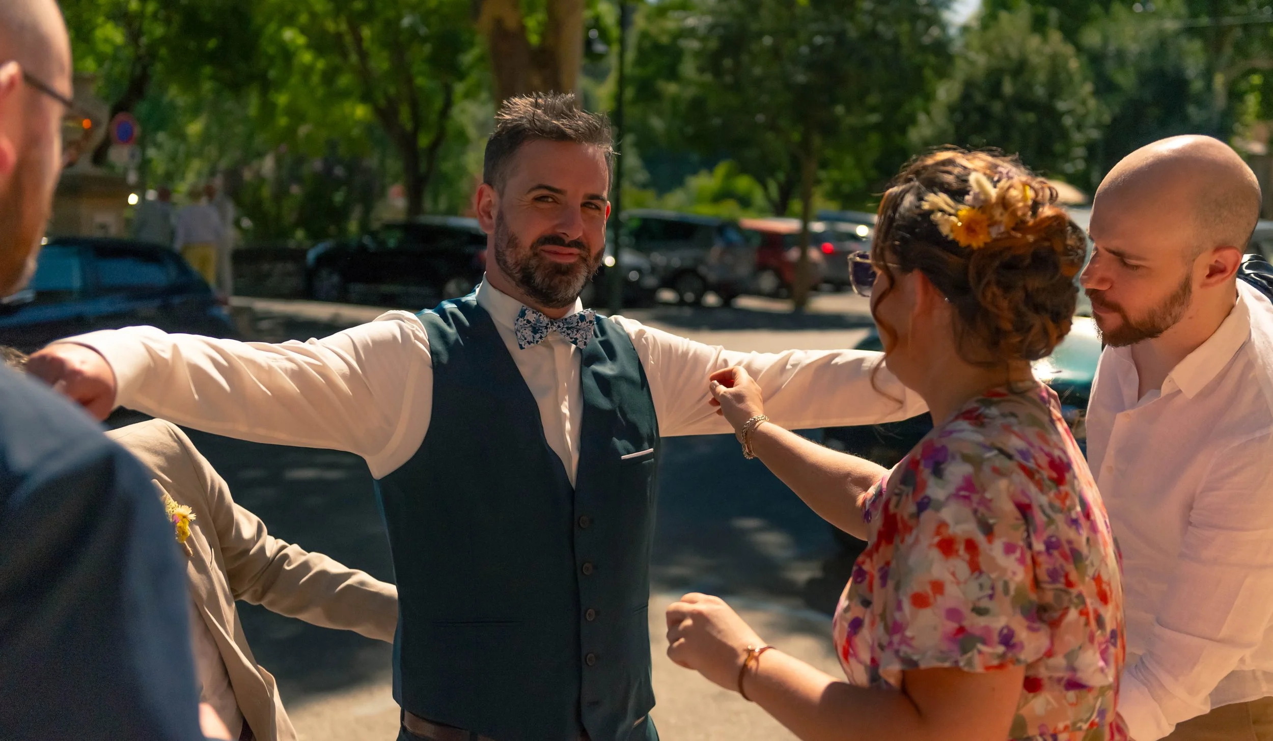 Groupe de personnes lors d'une célébration en plein air, un homme souriant au centre, portant une veste sans manches, une chemise à manches longues, un nœud papillon floral, entouré de 3 autres personnes.
