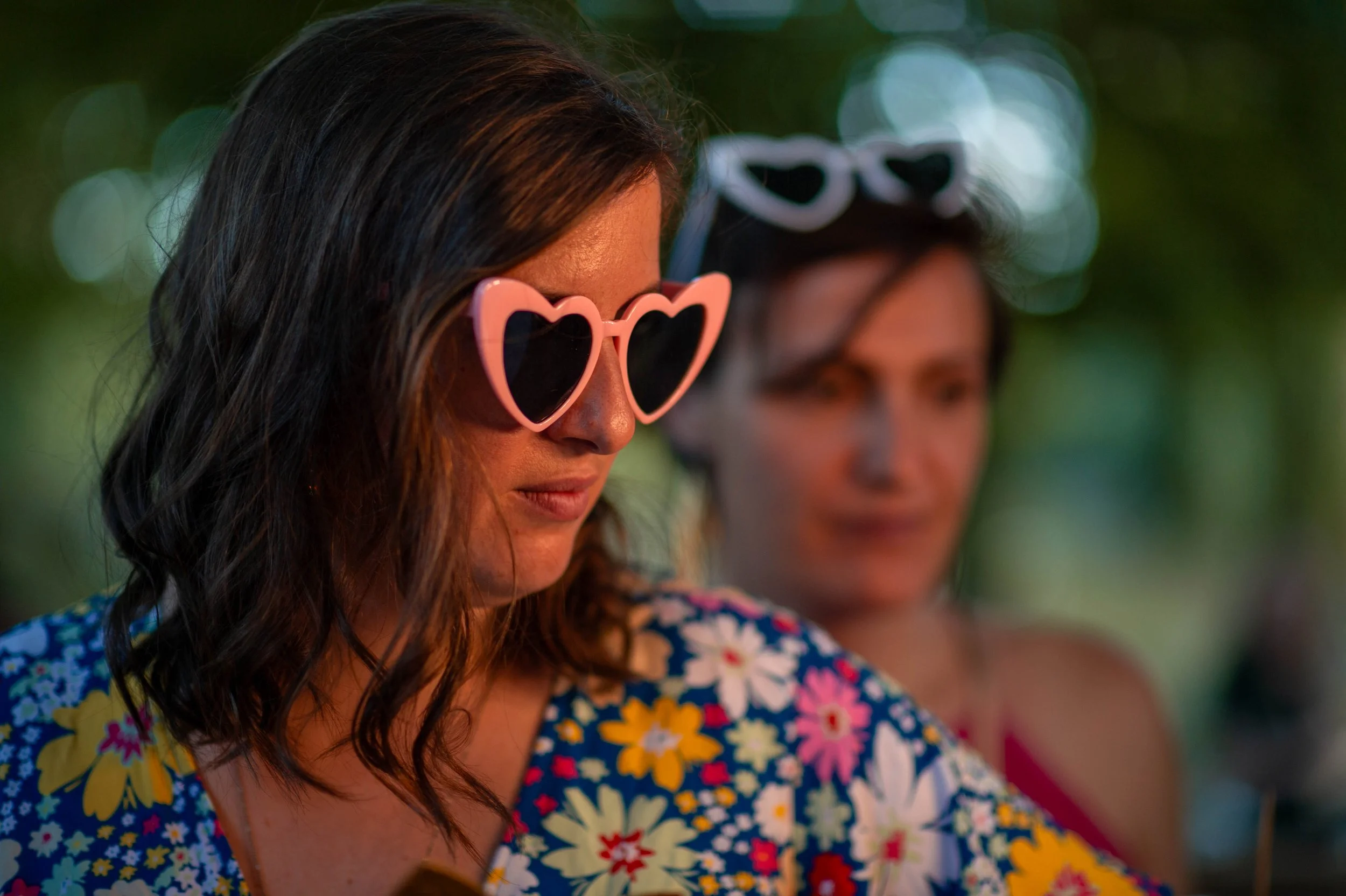 Deux femmes portant des lunettes de soleil en forme de cœur, l'une rose et l'autre blanche, lors d'une journée en plein air avec un fond flou de nature.