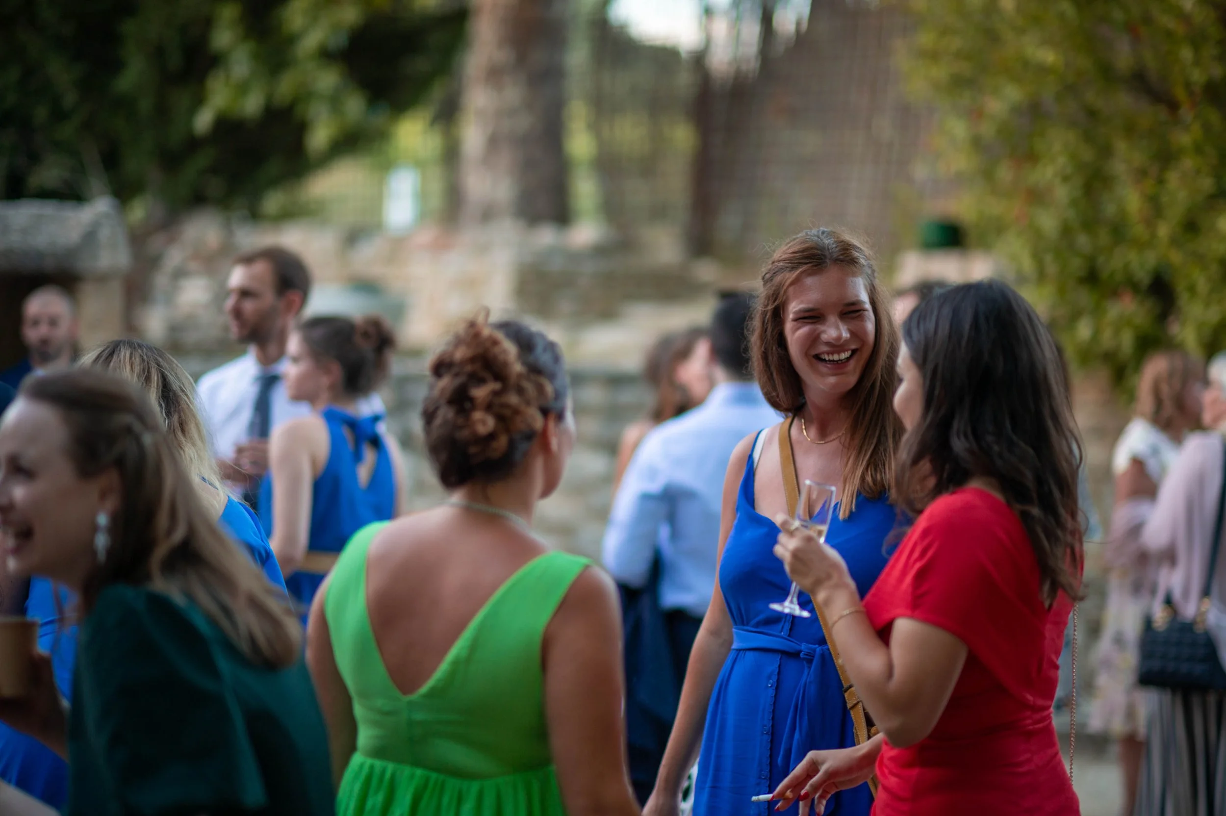 Groupe de personnes en fête à l'extérieur, souriantes, discutant, habillées en vêtements colorés, arrière-plan avec arbres et un mur en pierre.