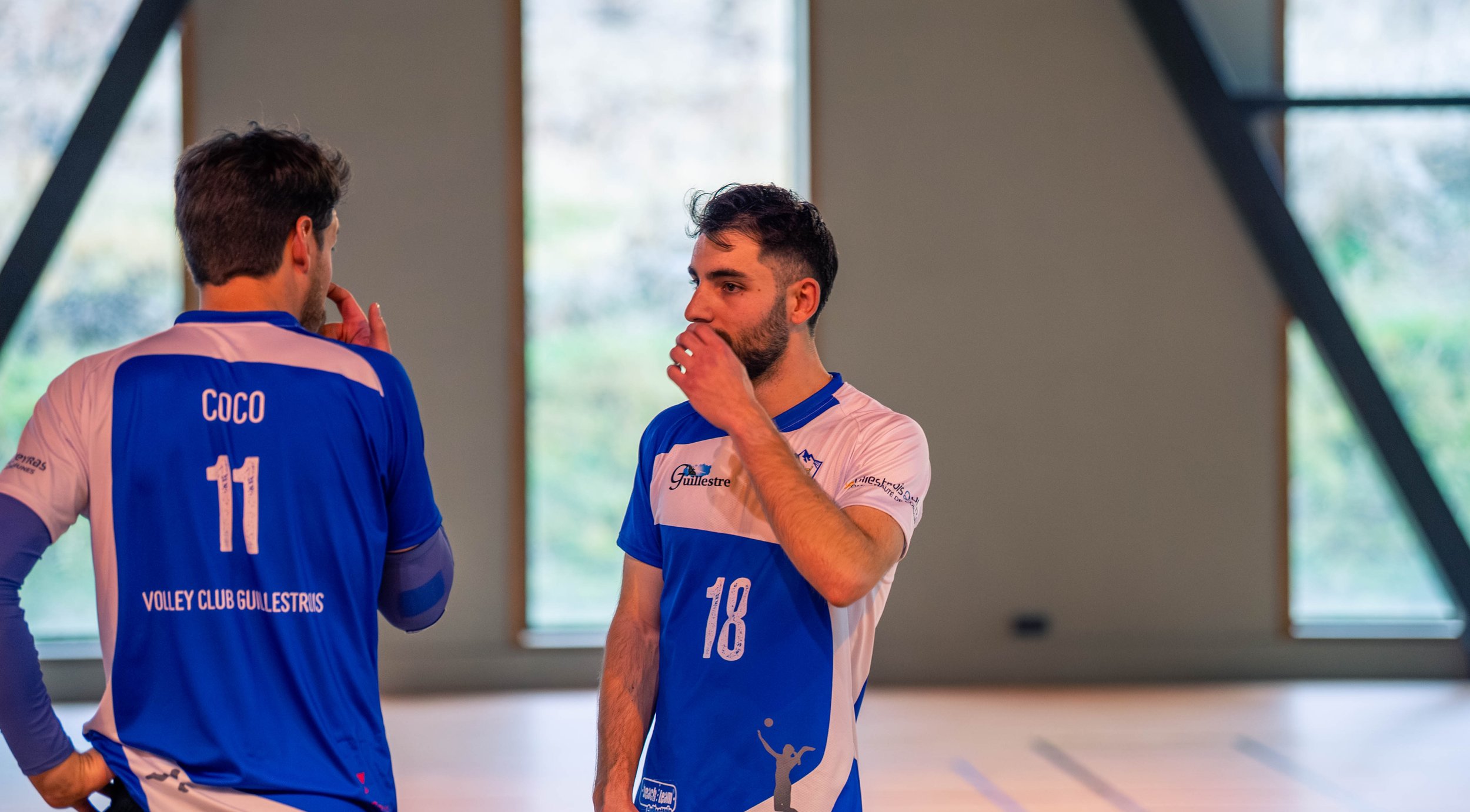Deux joueurs de volley-ball en conversation dans une salle d'entraînement, portant des maillots bleus et blancs. Un porte le numéro 11 avec le nom 'Coco', l'autre le numéro 18.