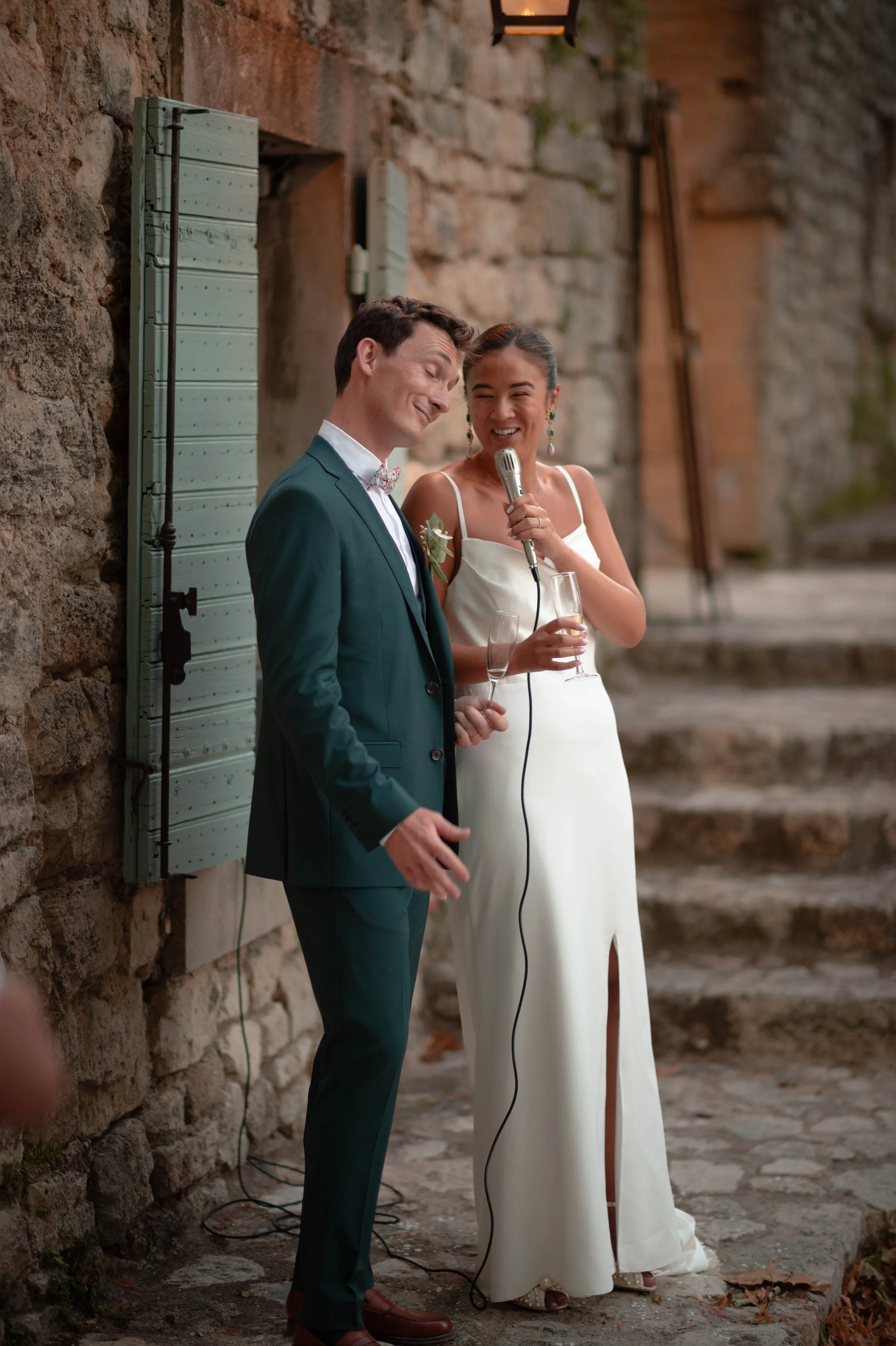 Un couple habillé pour un mariage, avec l'homme en costume bleu et la femme en robe blanche, lors d'une cérémonie en extérieur, la femme parlant dans un micro et tenant un verre de champagne.