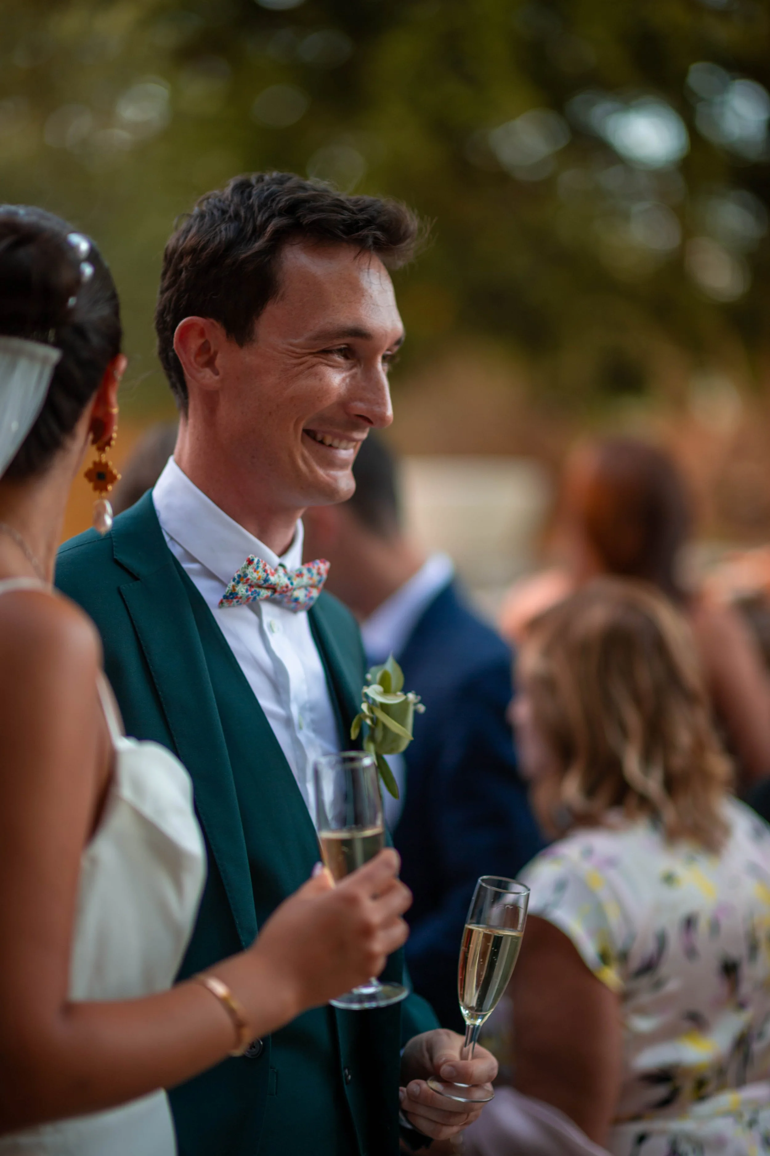Un homme souriant lors d'une réception, tenant une flûte de champagne, vêtu d'un costume vert et d'un nœud papillon coloré, entouré d'autres personnes en tenue élégante.