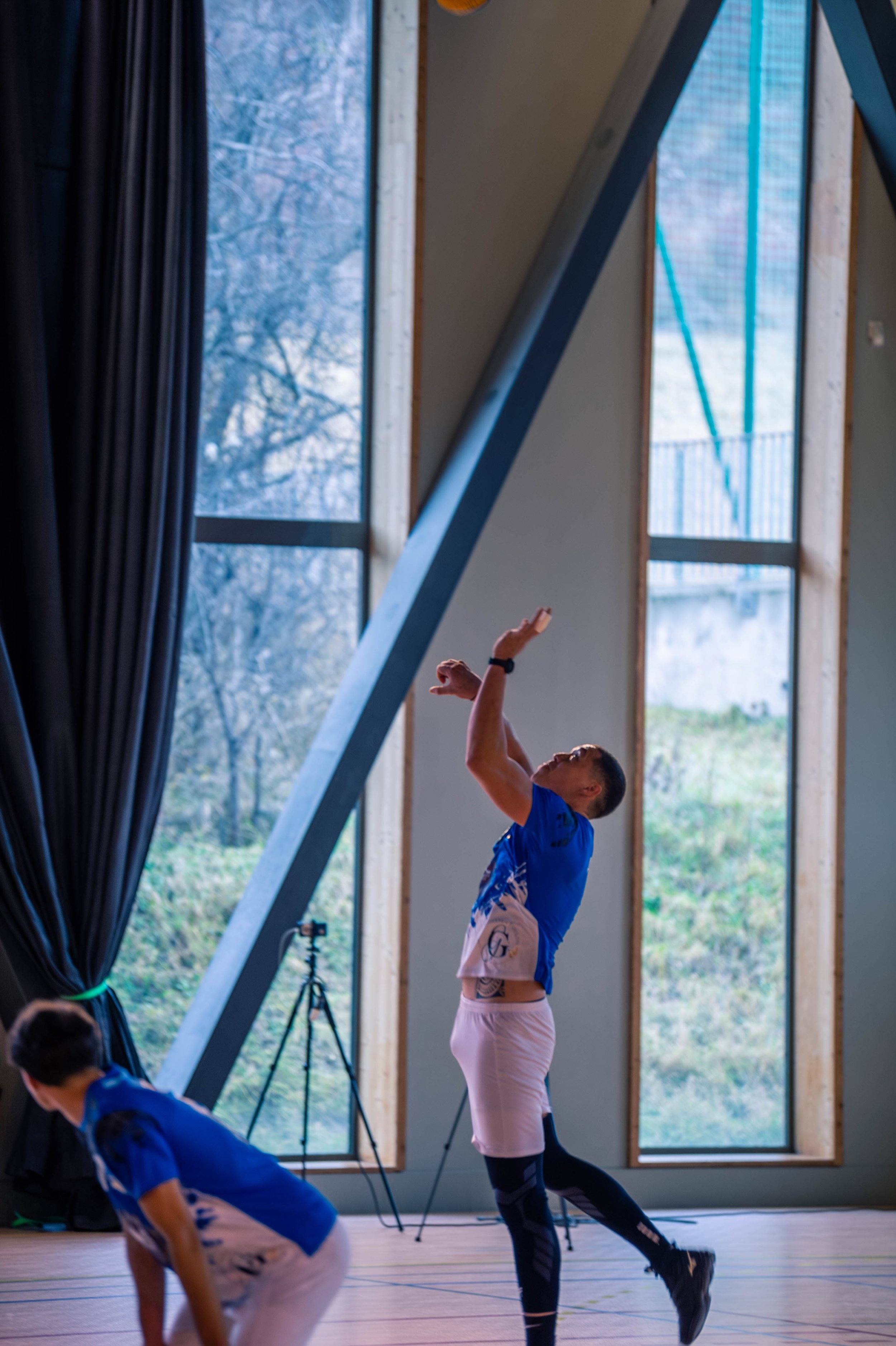 Deux athlètes en vêtements bleus pratiquant le volleyball dans un gymnase avec de grandes fenêtres et des arbres visibles à l'extérieur.