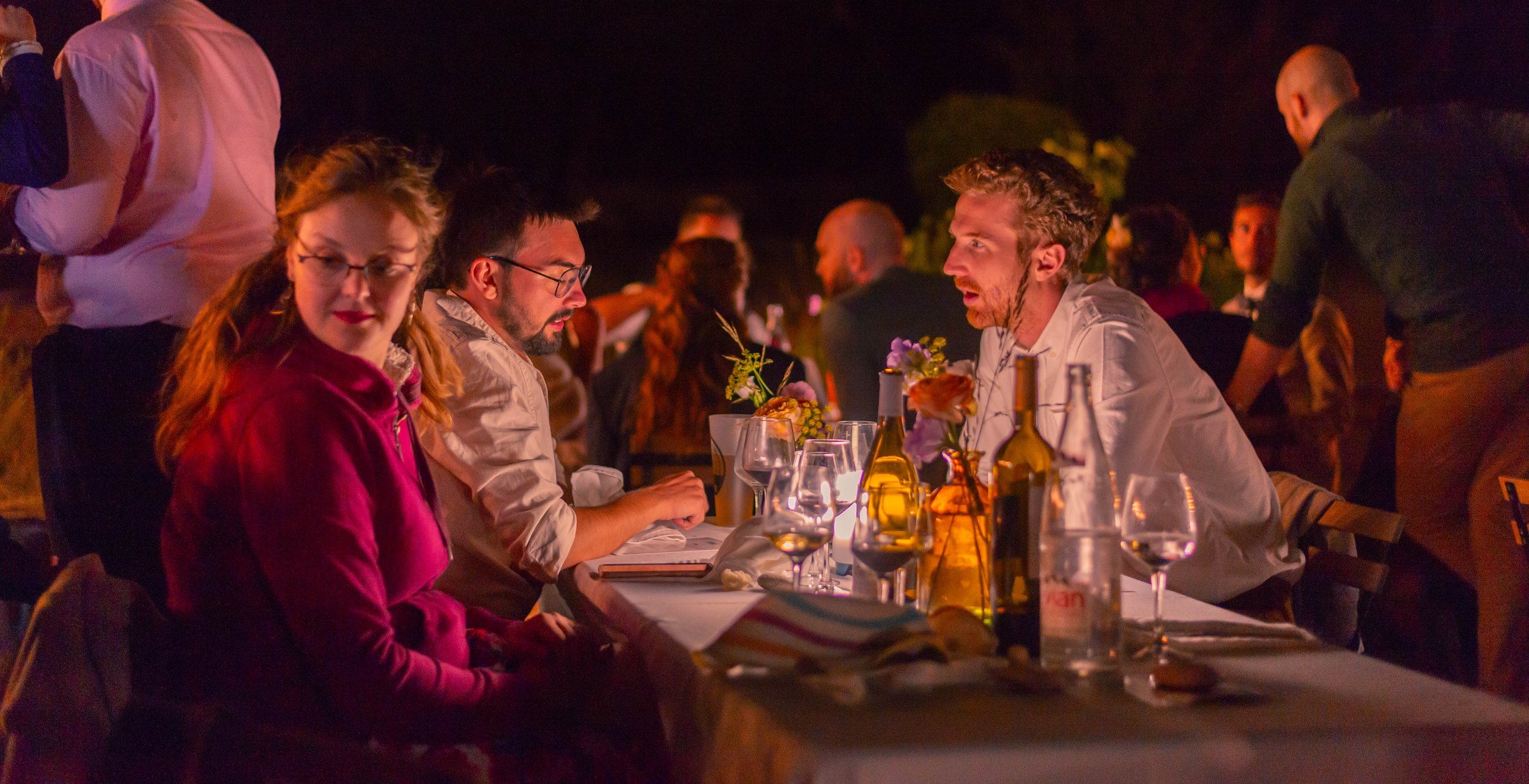 Groupe de personnes lors d'un dîner en plein air la nuit, assises autour d'une table avec des bouteilles et des fleurs, ambiance chaleureuse et conviviale.