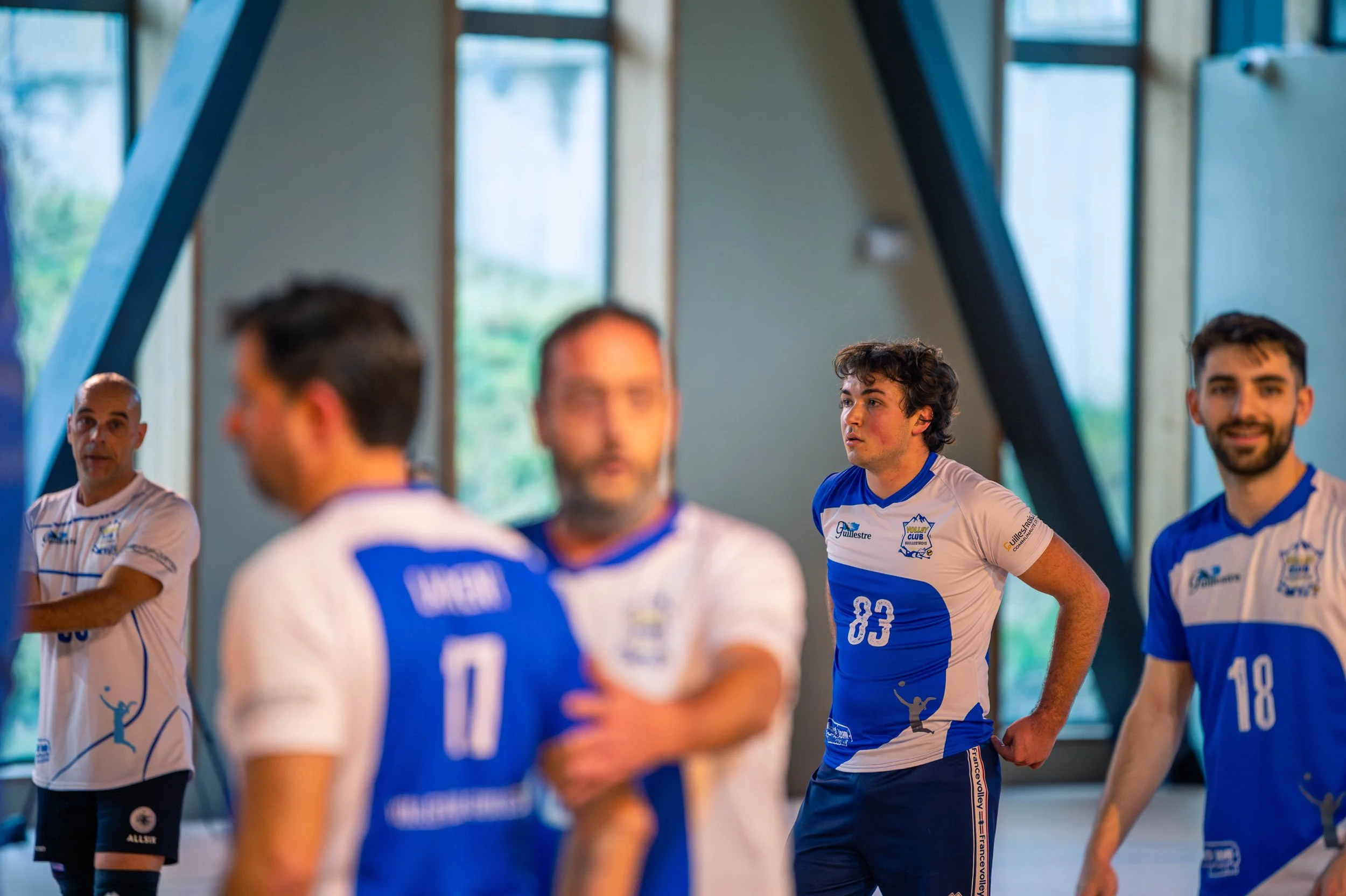 Joueurs de volleyball en tenue bleue et blanche dans une salle intérieure, discutant ou préparant leur jeu.