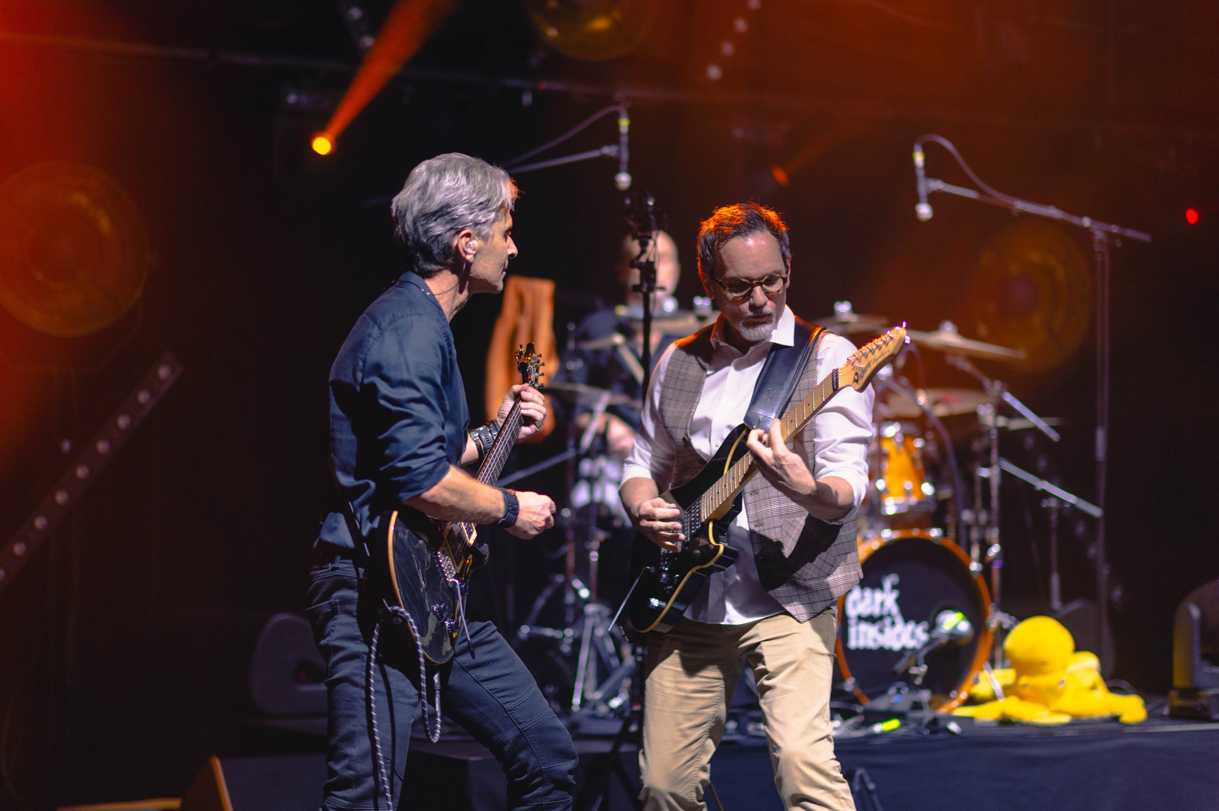 Deux musiciens jouent de la guitare sur scène avec un batteur en arrière-plan, sous un éclairage orange et jaune.