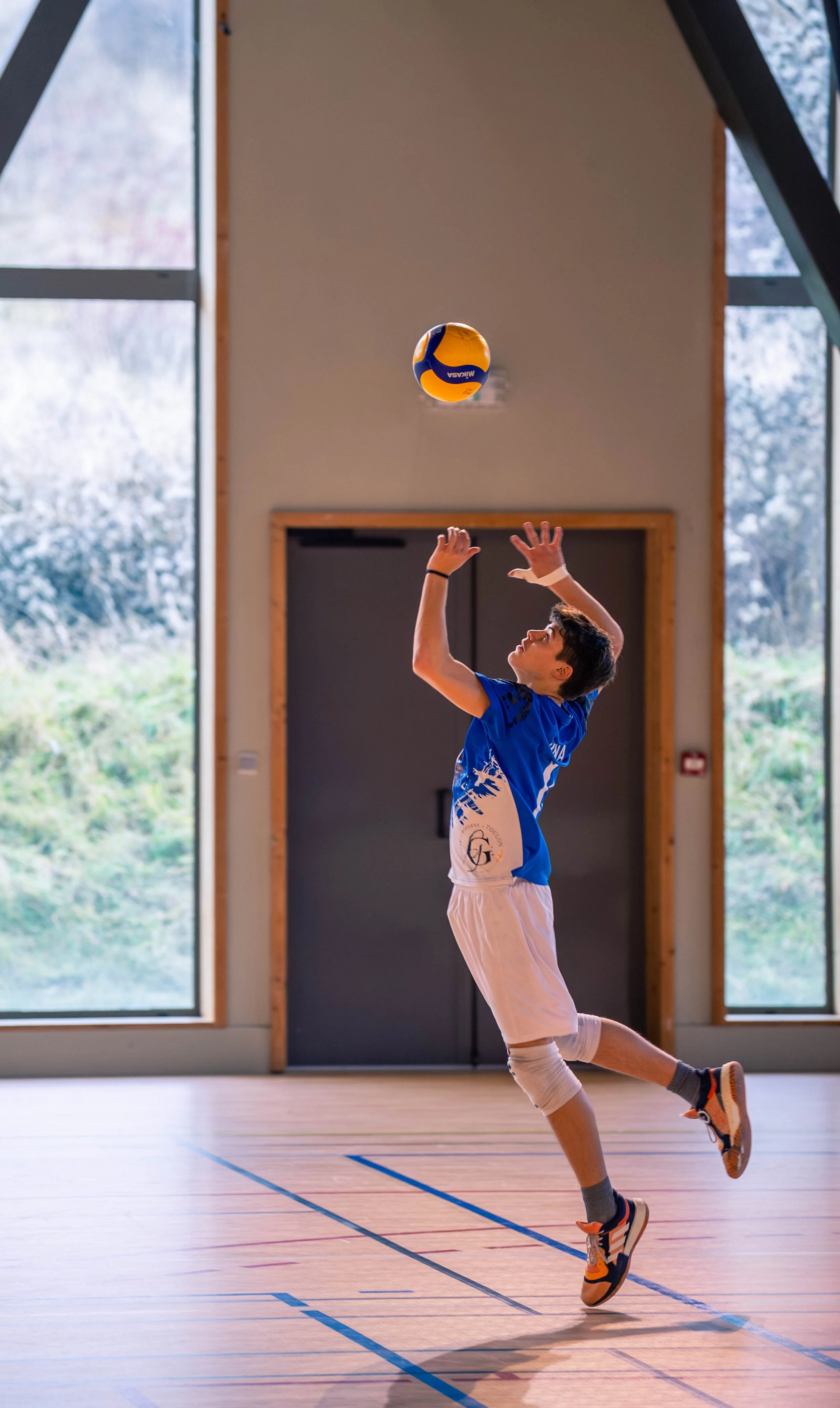 Un jeune garçon jouant au volley-ball à l'intérieur d'un gymnase, prêt à servir le ballon, portant un maillot bleu, un short blanc et des chaussures de sport.