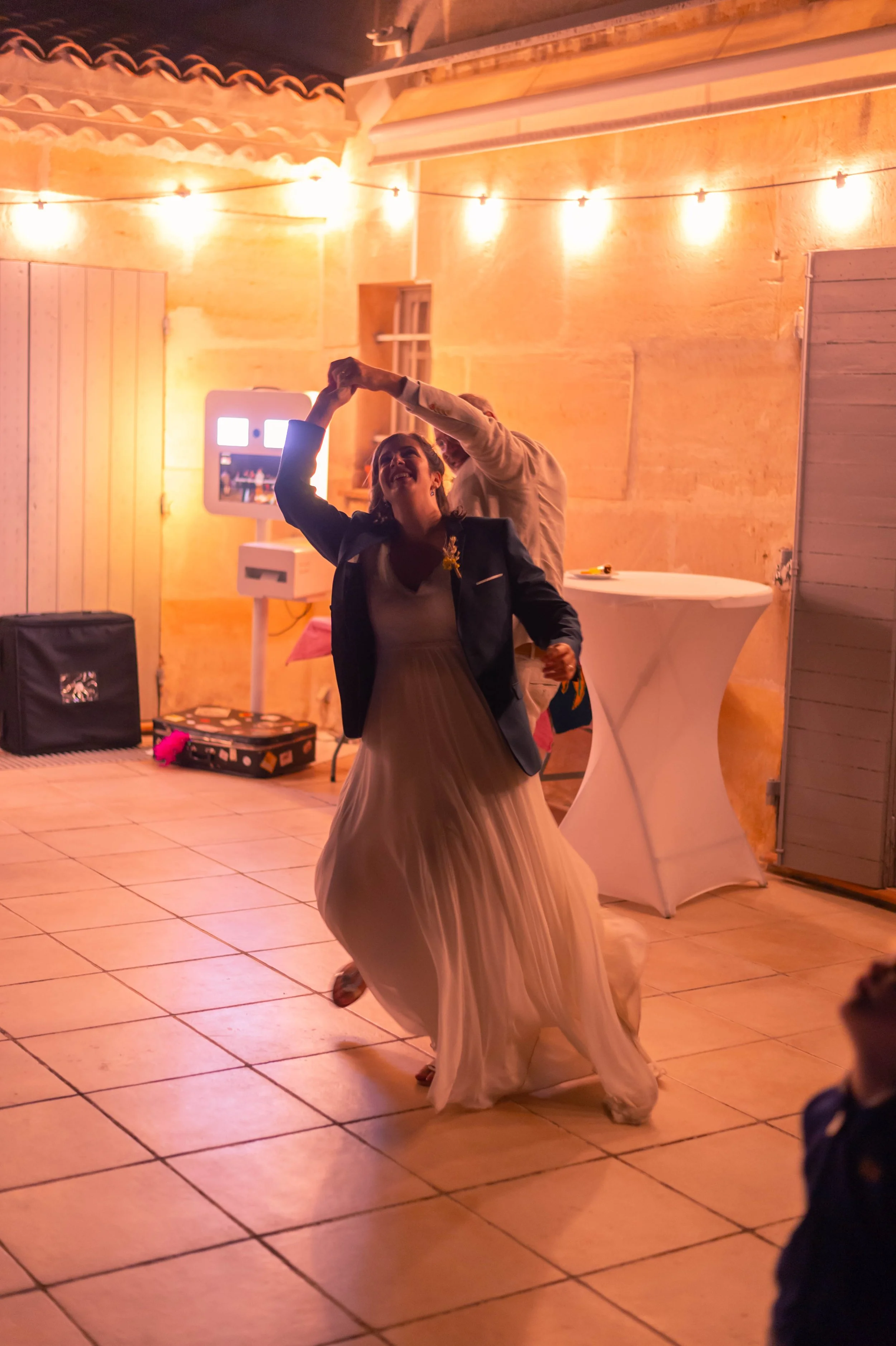Une femme danse lors d'une fête, portant une robe longue blanche et une veste sombre, avec un homme derrière elle dans une ambiance chaleureuse et éclairée par des guirlandes lumineuses.