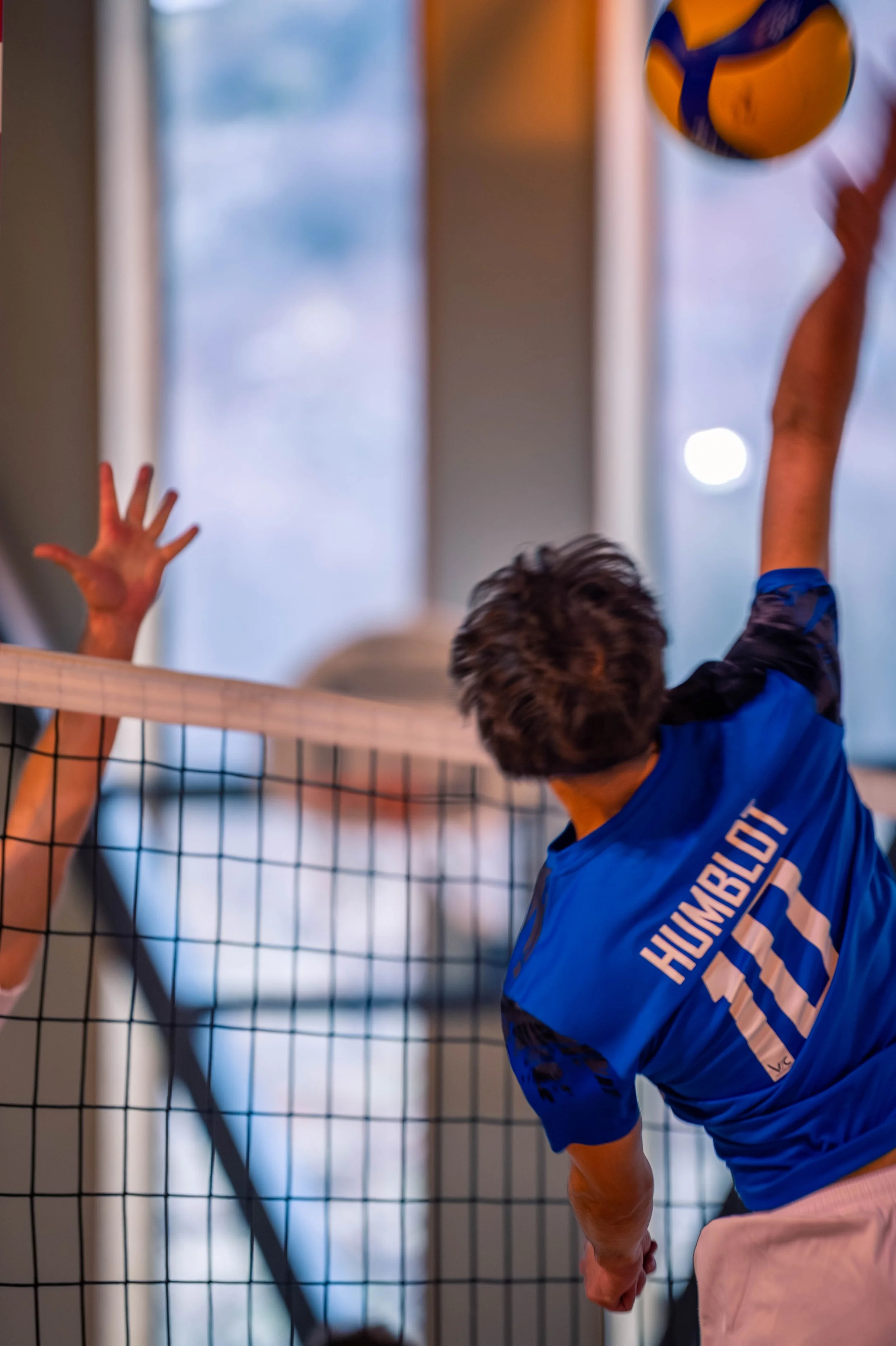 Joueur de volley-ball en action, sautant pour attaquer ou bloquer, avec un adversaire dont la main est levée pour contrer, dans une salle d'intérieur.