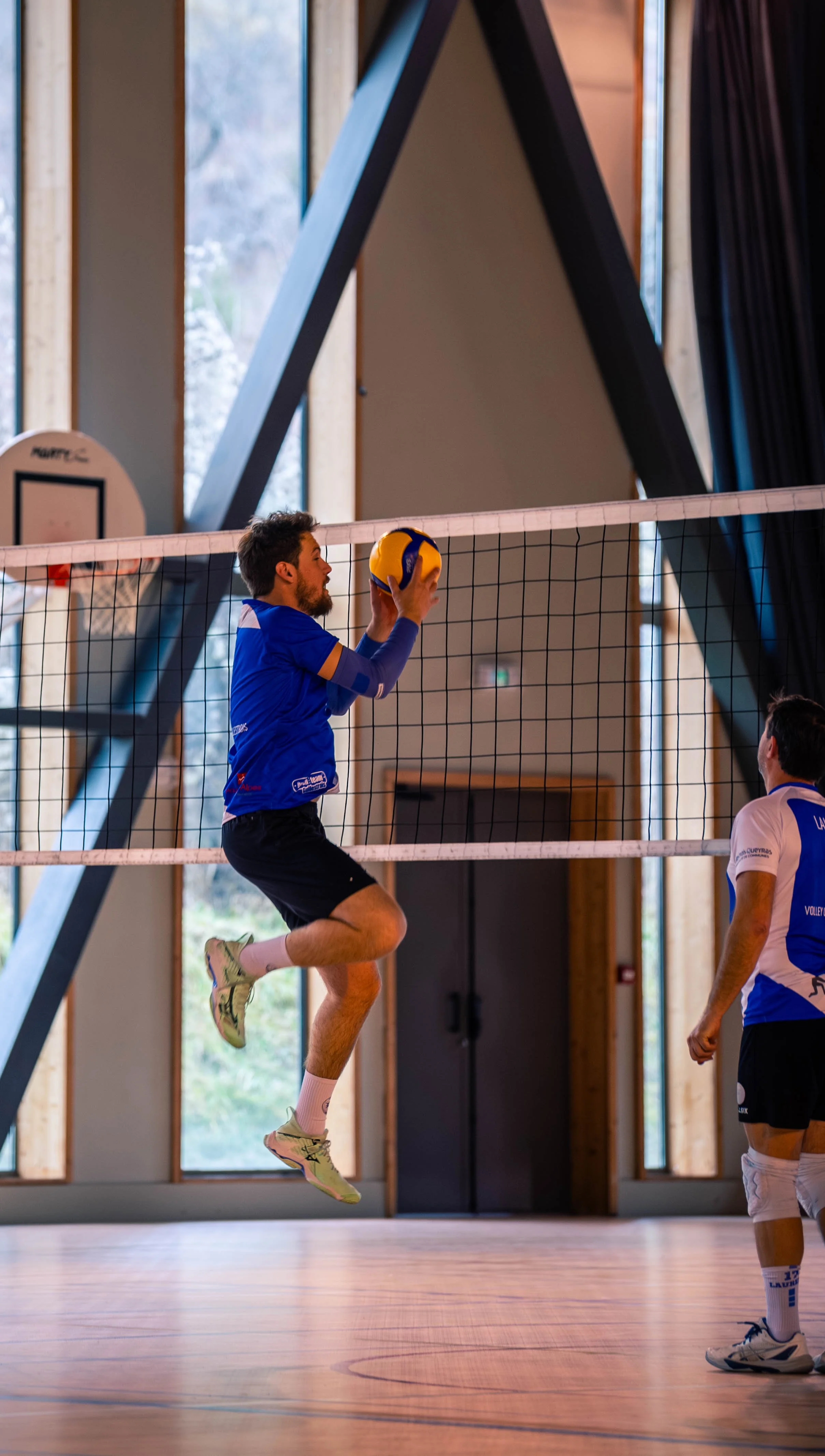Deux joueurs de volleyball en action dans un gymnase, one sautant pour attaquer le ballon, les autres observant