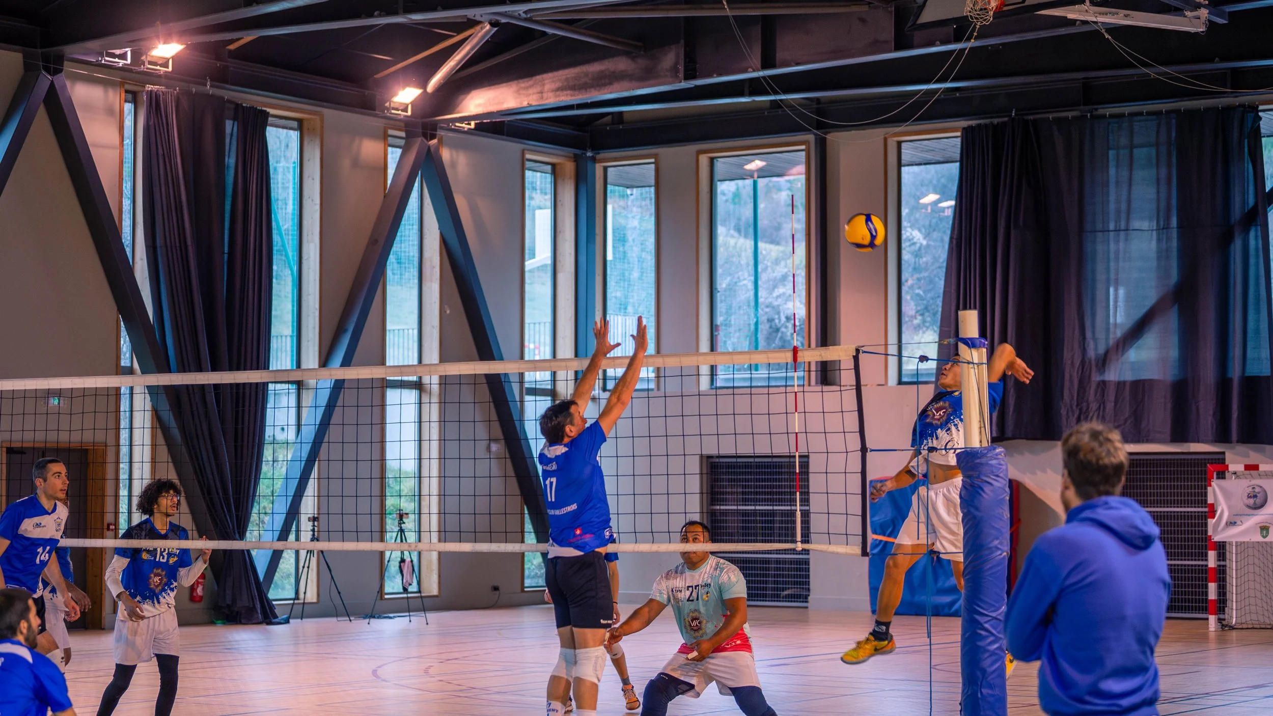 Une scène dvolley-ball en intérieur, avec un joueur en noir et bleu sautant pour attaquer le ballon, tandis qu'un défenseur en blanc et bleu tente de bloquer, d'autres joueurs et un entraîneur observant autour.