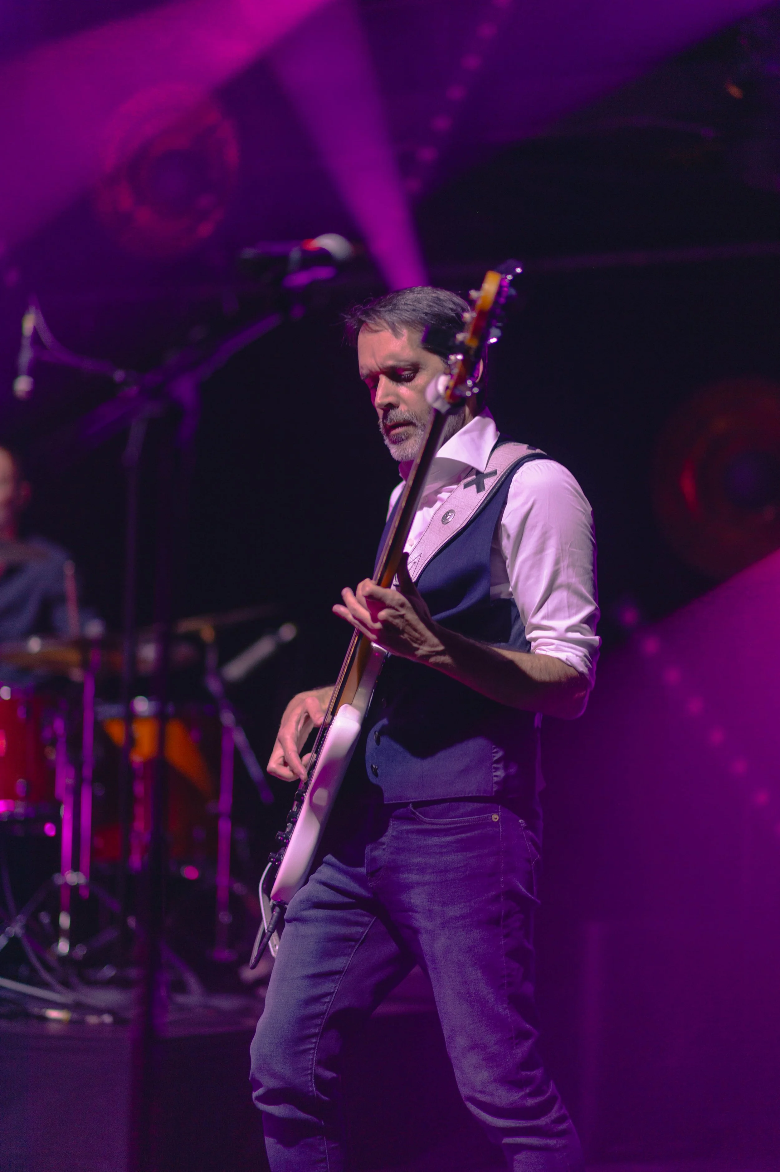 Un homme jouant de la guitare lors d'un concert, éclairé par des lumières violettes et roses, avec un clavier visible en arrière-plan.