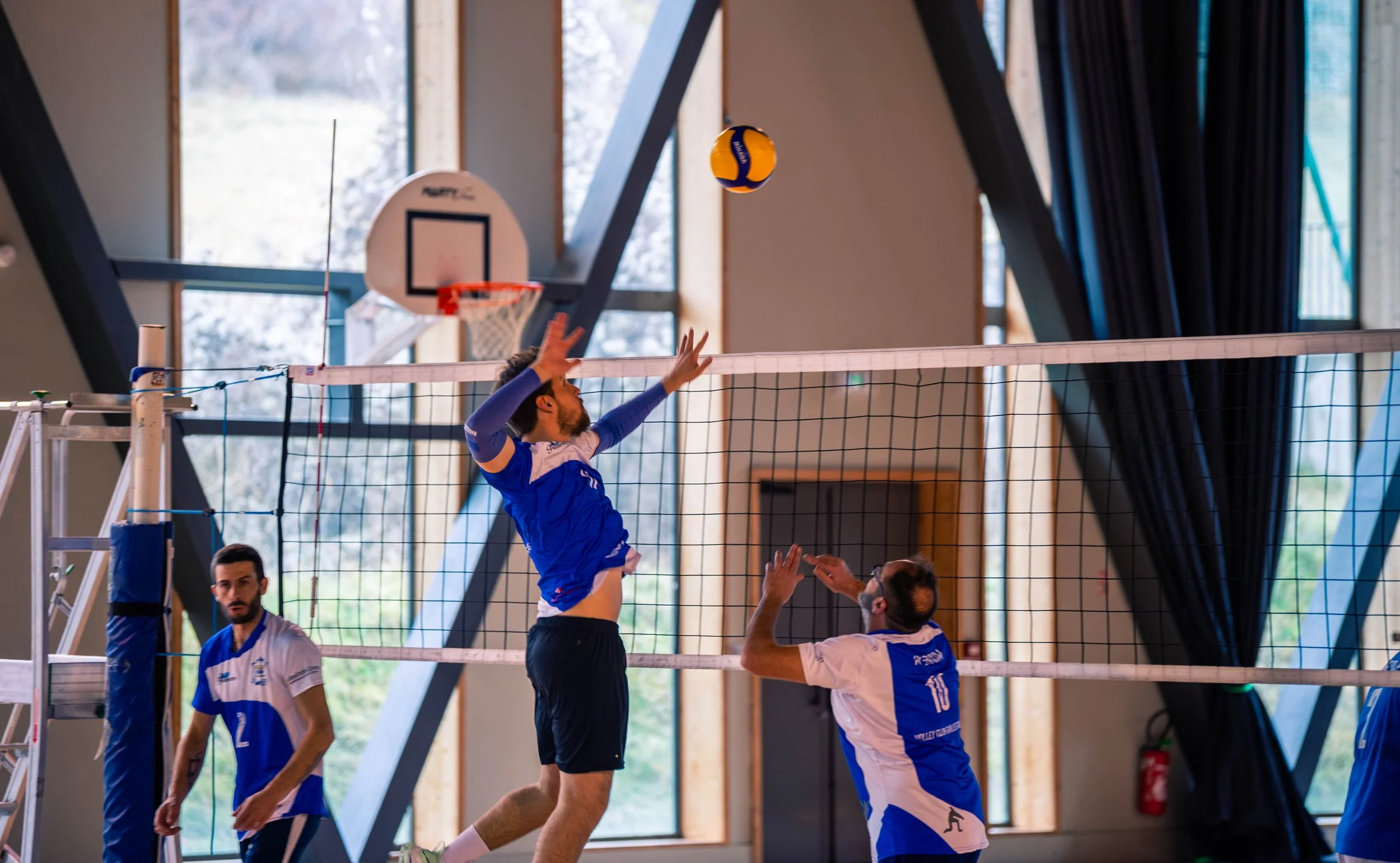 Une équipe de volley-ball en action lors d'un match intérieur, avec un joueur en train de sauter pour attaquer la balle, un autre prêt à réceptionner, dans un gymnase avec des fenêtres en arrière-plan.