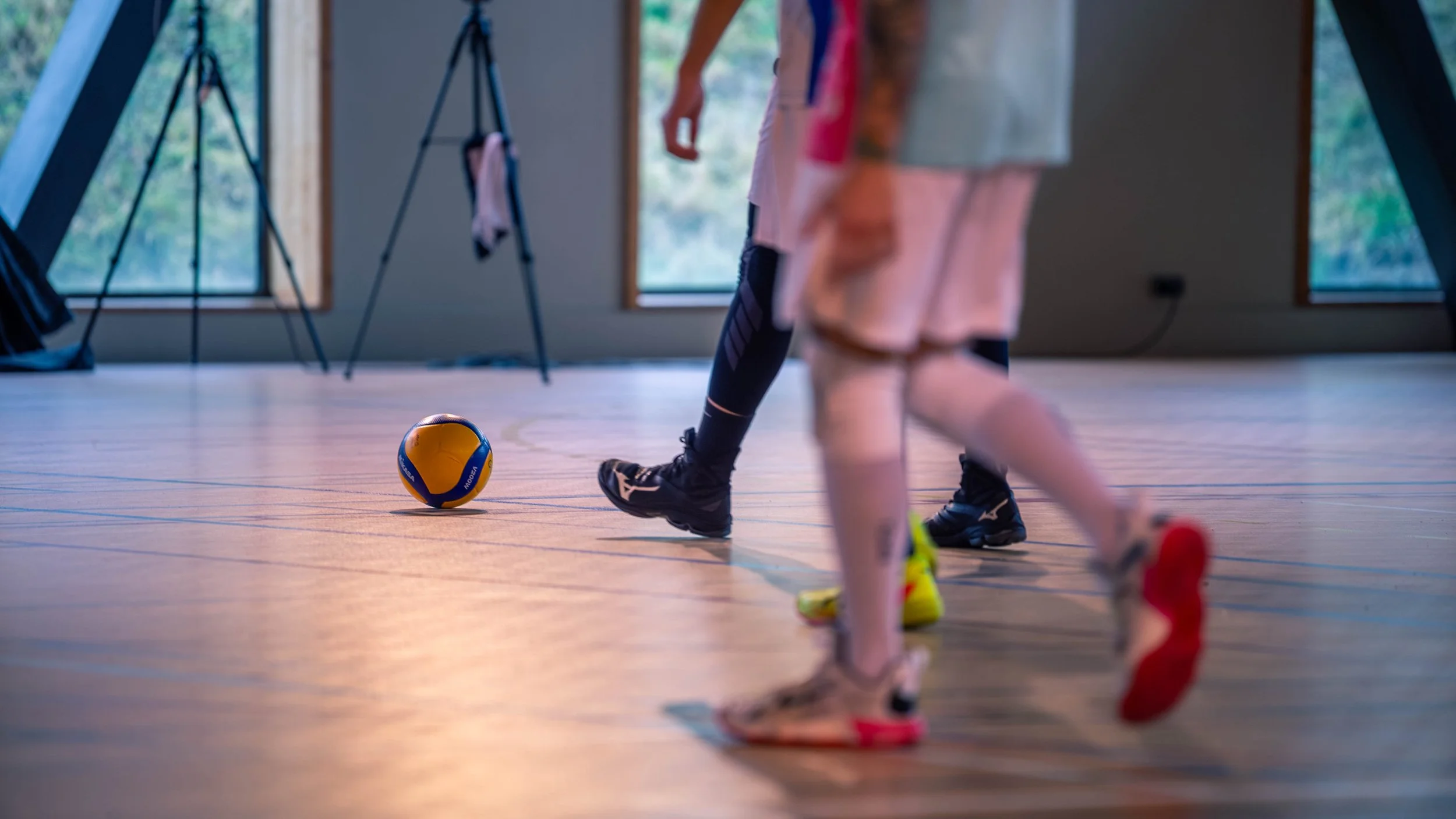 Entraînement de volleyball en intérieur, vue des jambes des joueurs avec un ballon évoqué sur le sol.