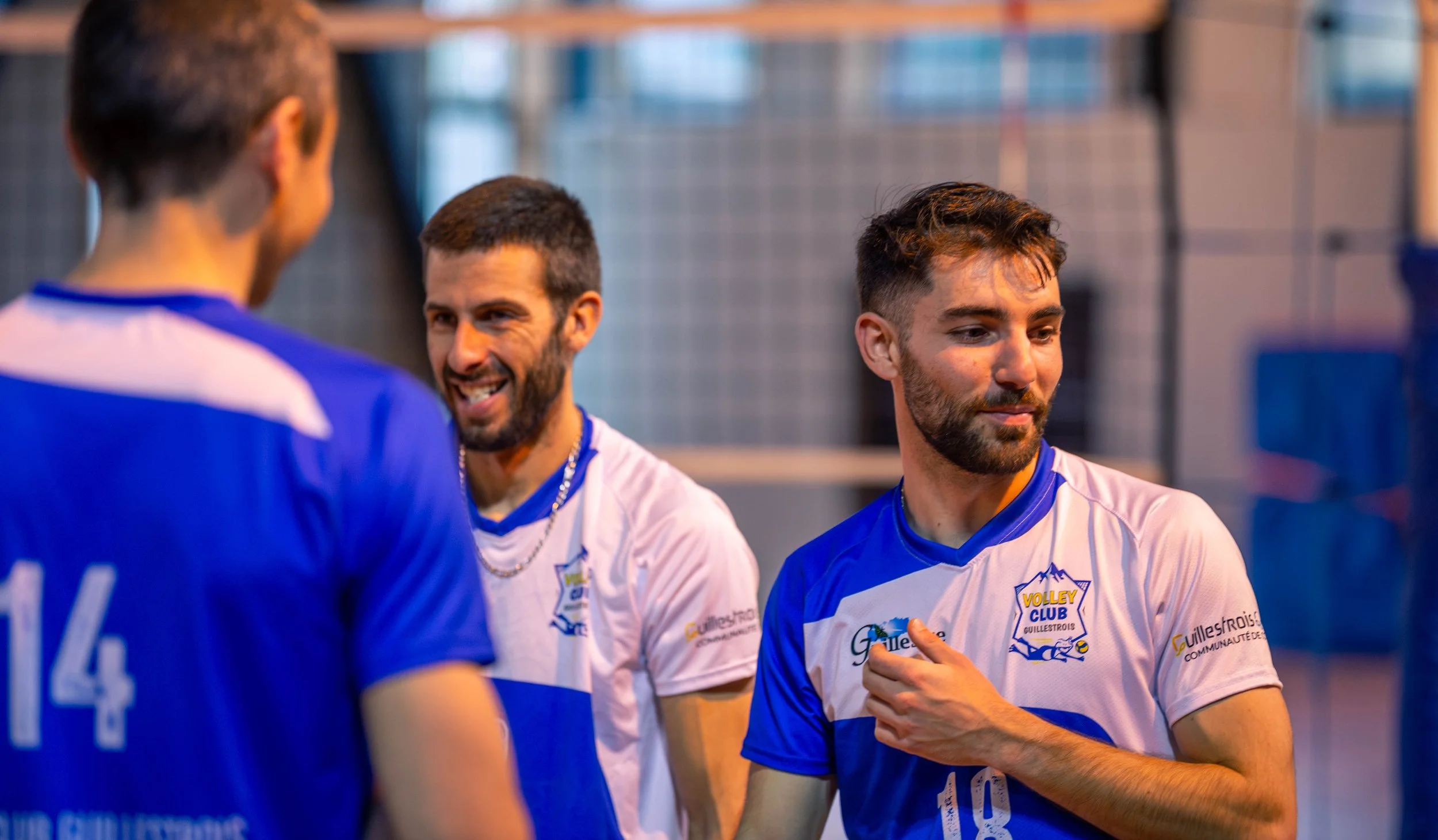 Trois joueurs de volley-ball discutent sur un terrain, portant des maillots bleus et blancs avec le logo du club Volley Club Guilléstrois.