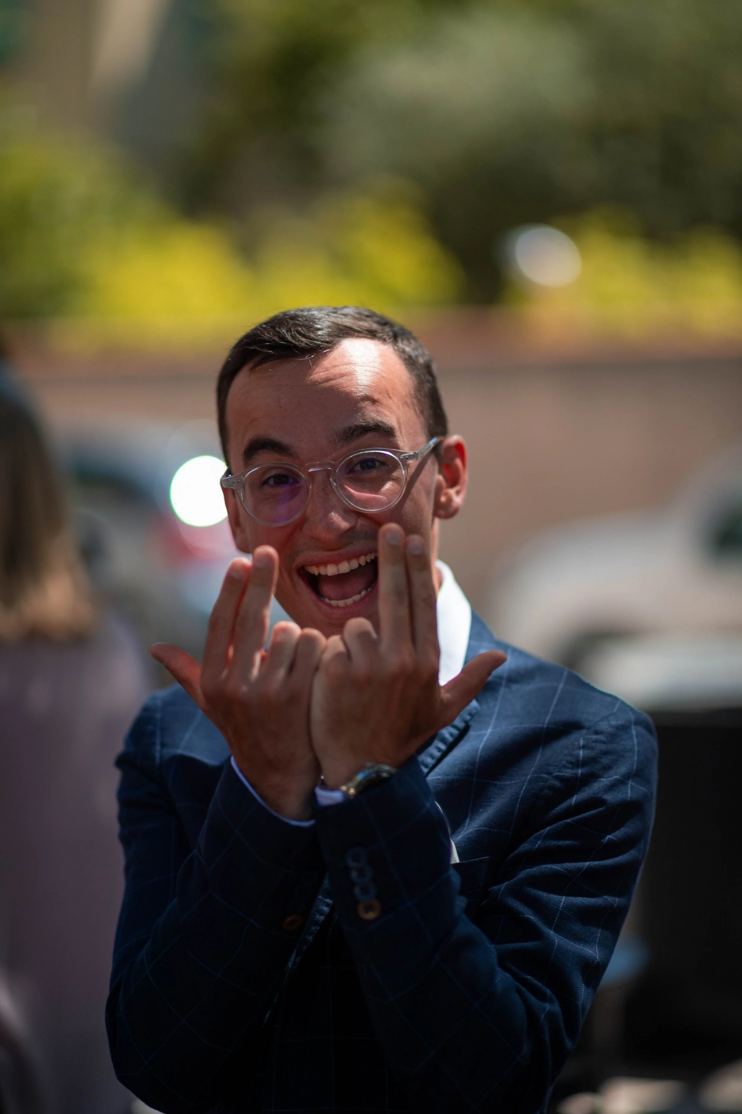 Un jeune homme souriant, portant des lunettes, en plein rire ou en joie, dans un environnement extérieur.