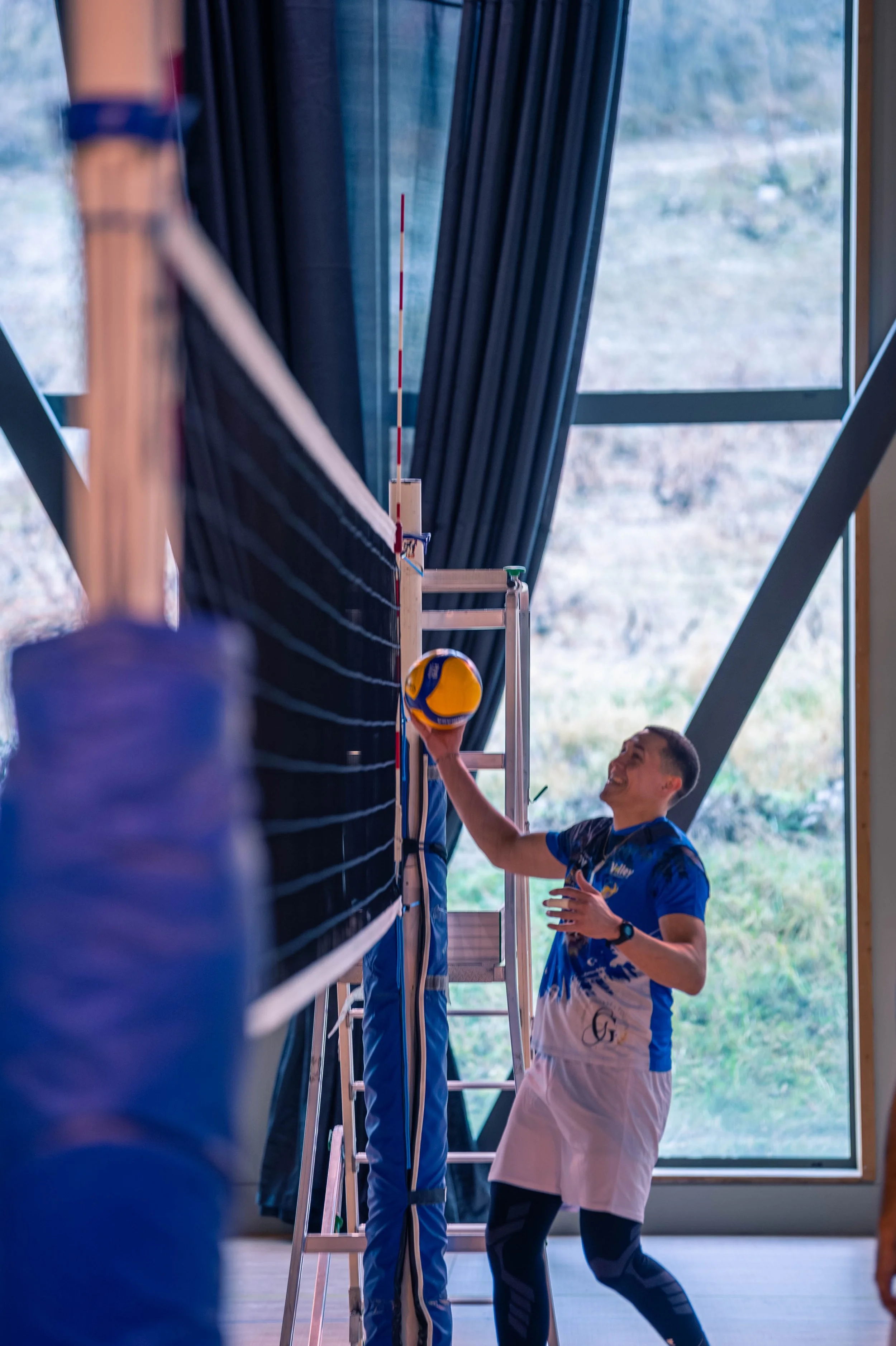 Un homme joue au volleyball à l'intérieur, il tient un ballon jaune et bleu, avec un filet de volleyball devant lui et une échelle à côté, dans une salle avec de grandes fenêtres en arrière-plan.