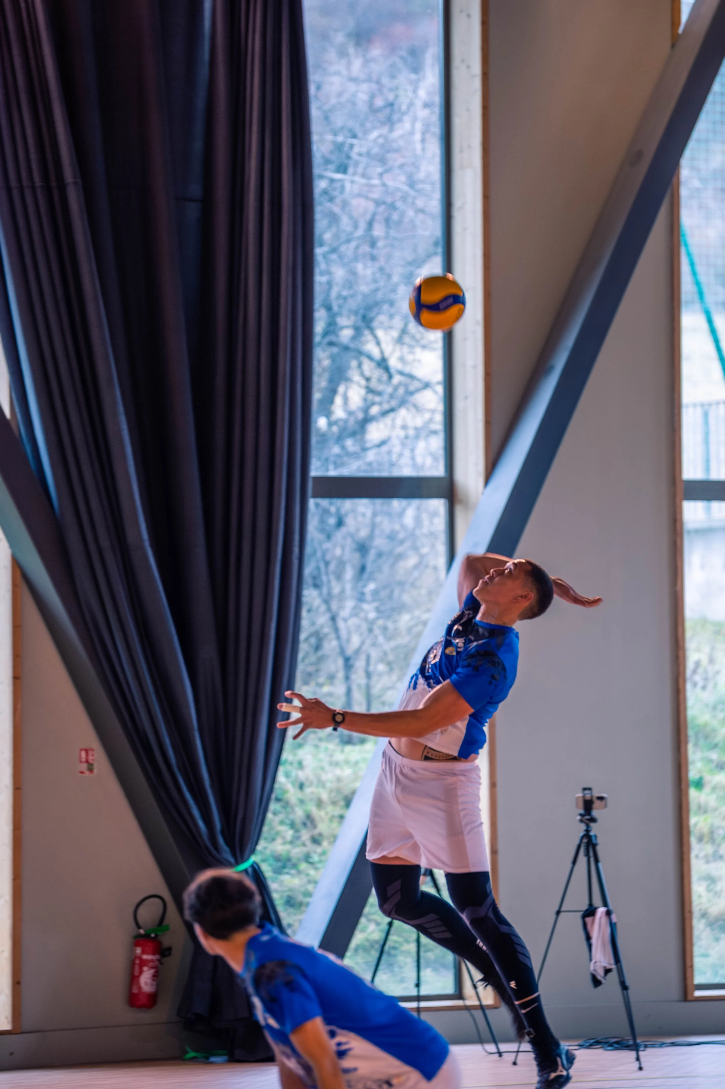 Deux joueurs de volleyball en action à l'intérieur, un homme le frappant avec la main tendue vers le ballon, un autre au sol, un appareil photo sur un trépied à côté.