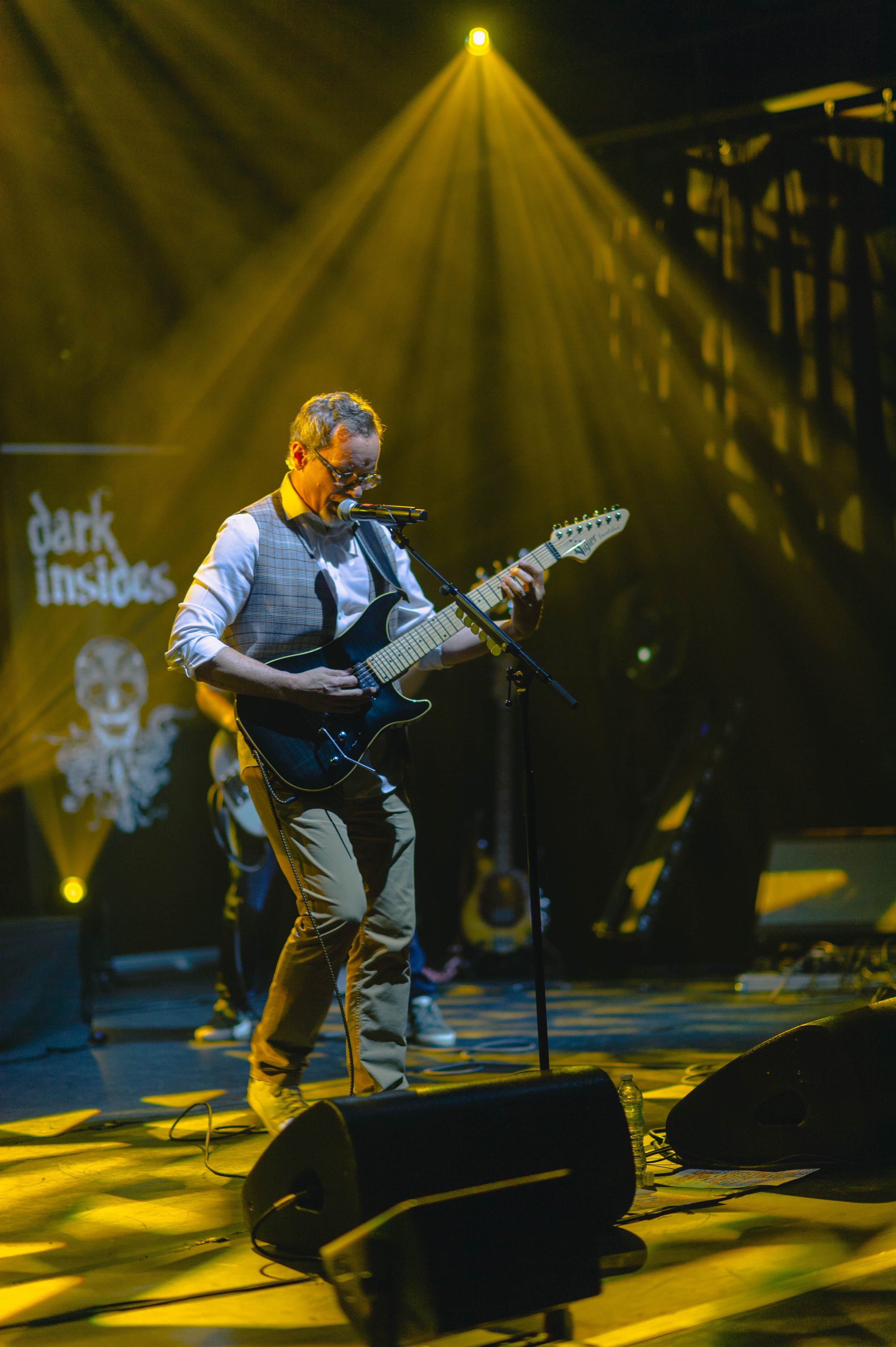 Musicien jouant de la guitare électrique en performace sur scène, éclairé par un éclairage jaune, avec un arrière-plan sombre et un logo 'dark insides'.