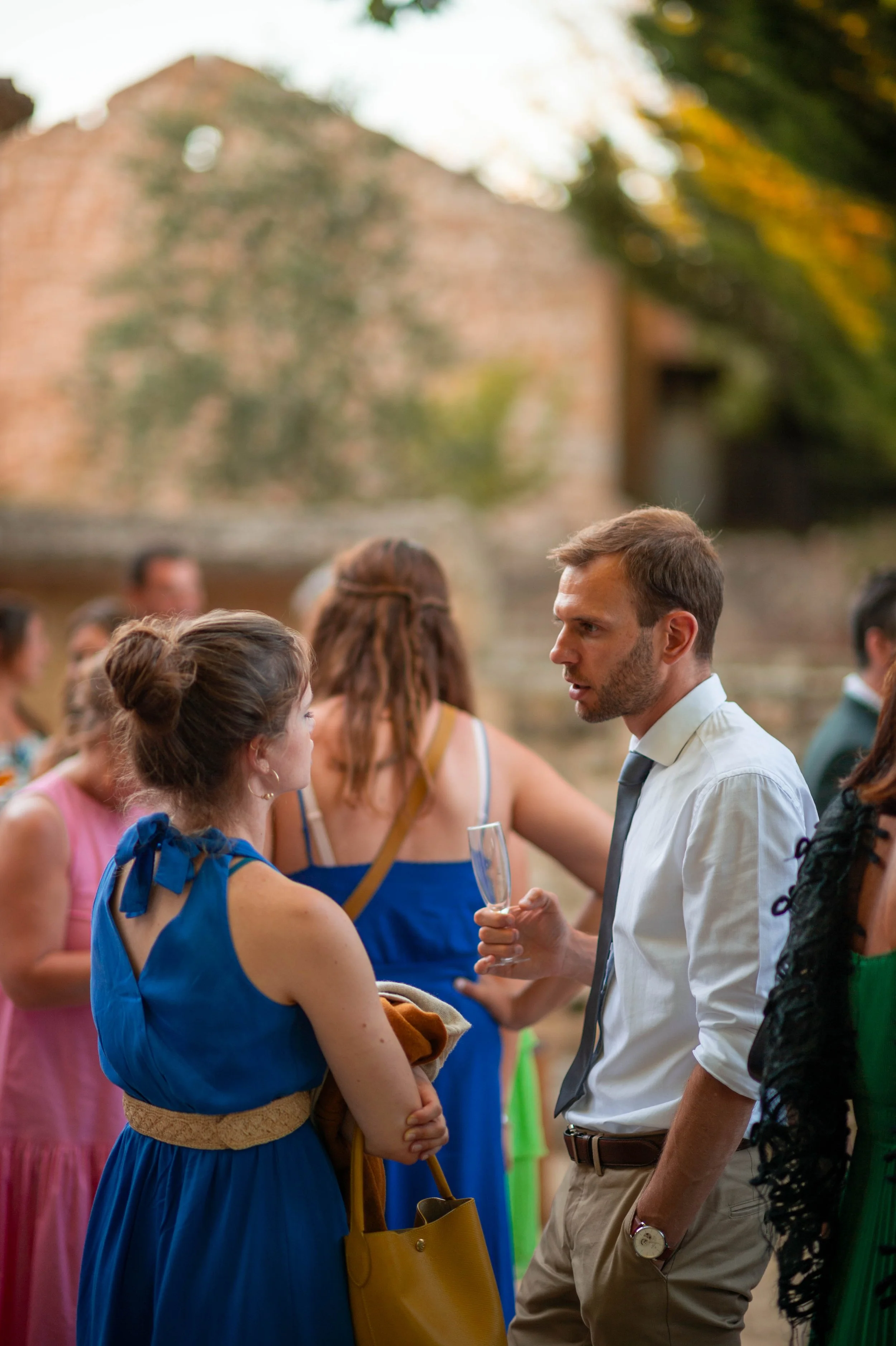 Un groupe de personnes lors d'une fête ou d'un rassemblement social en plein air, avec un homme en chemise blanche et cravate discutant avec une femme en robe bleue, parmi d'autres invités.