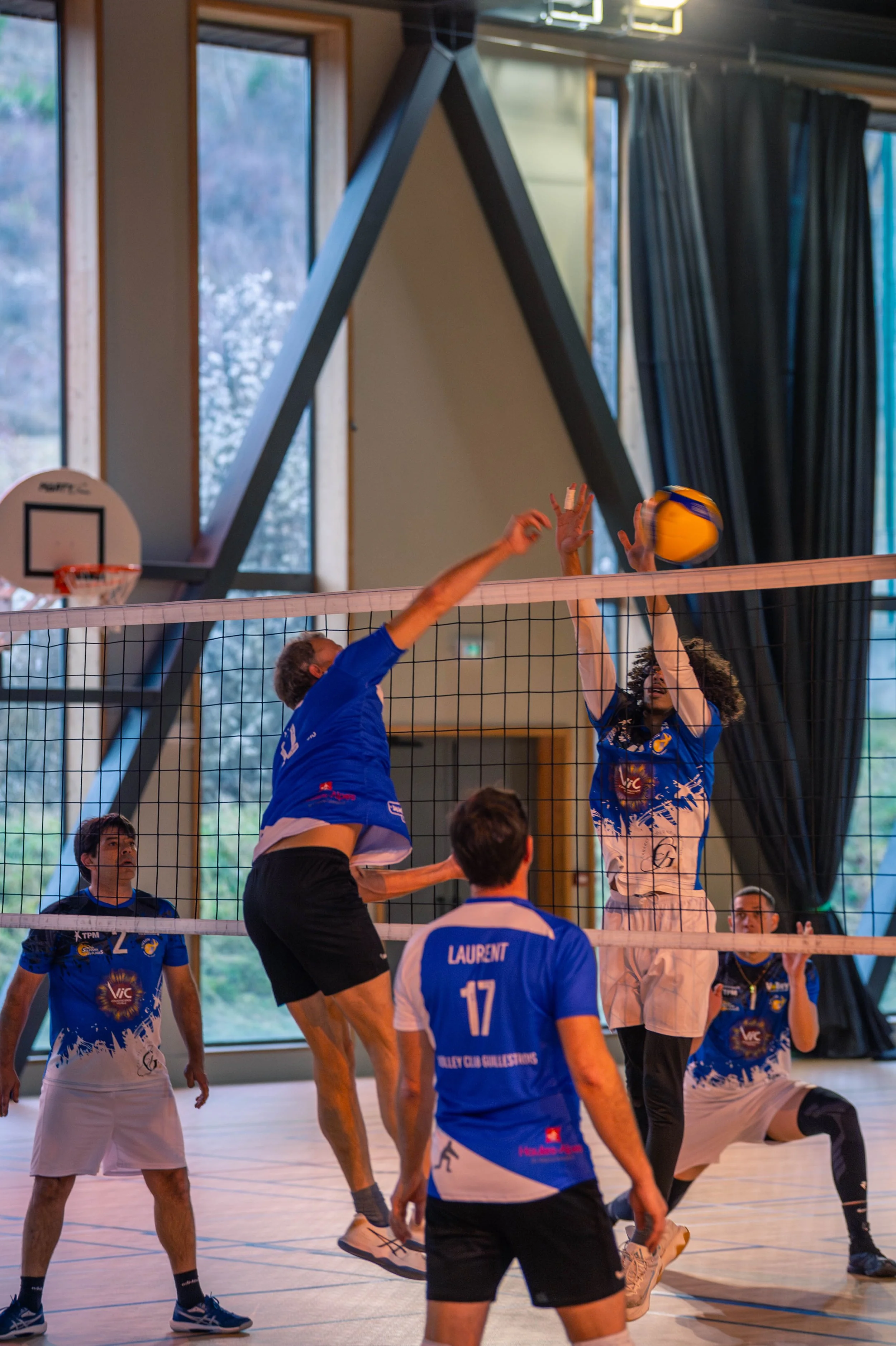 Une scène de volleyball en intérieur avec six joueurs, trois en bleu et blanc, un en noir, une femme avec des cheveux bouclés, en train de sauter pour bloquer la balle, et deux autres observant la partie.