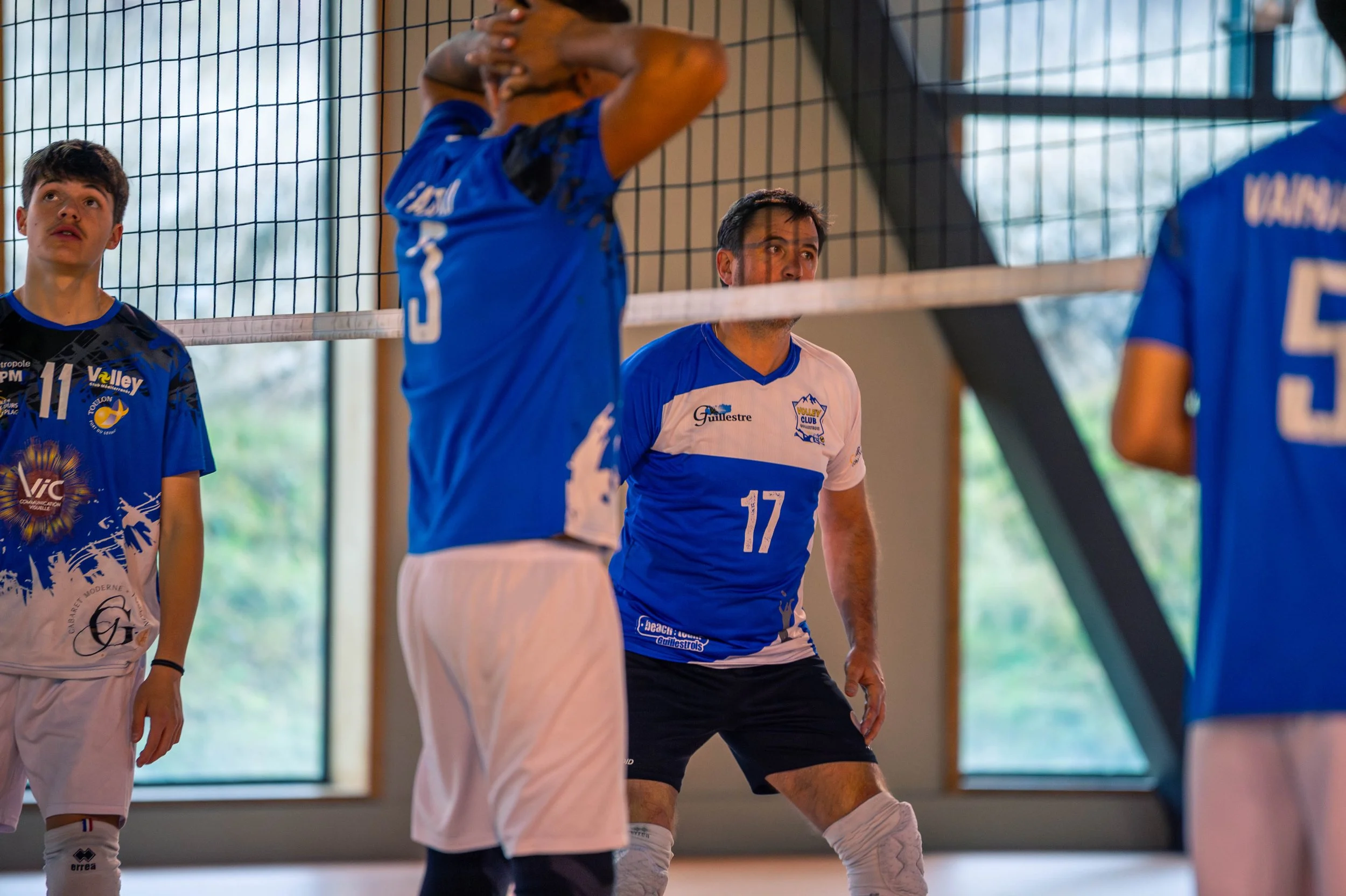 Joueurs de volley-ball en position d'attente lors d'un match en intérieur, portant des maillots bleus avec des numéros, derrière un filet de volley.