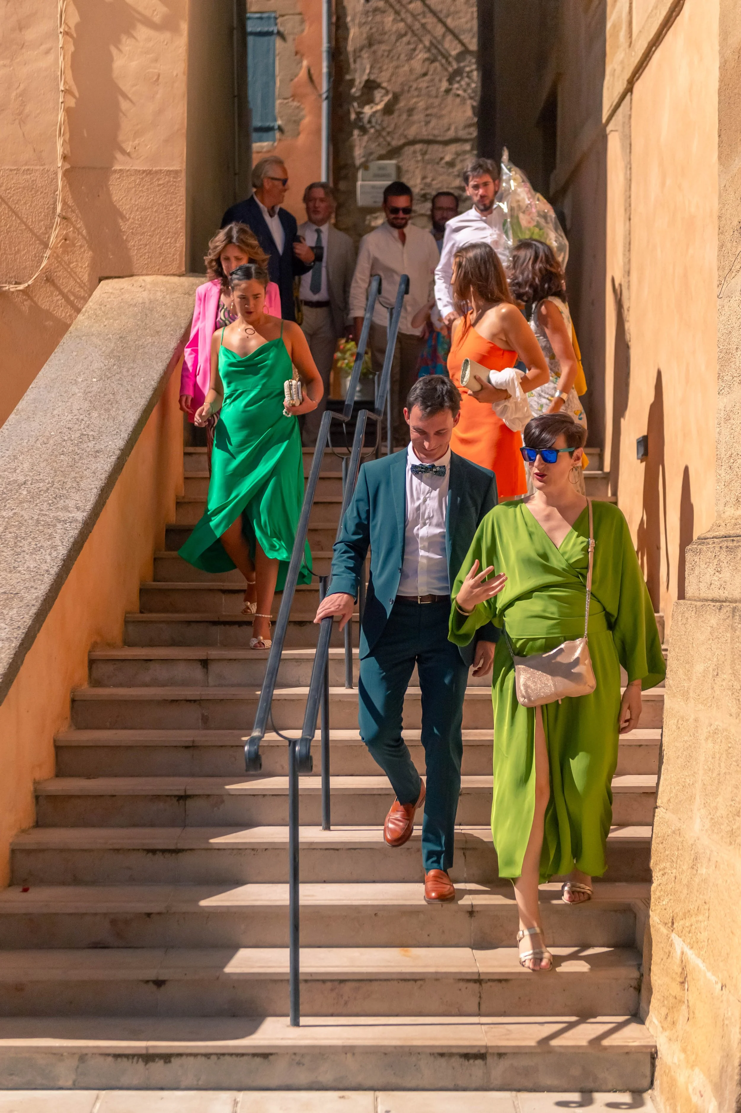 Groupe de personnes élégamment habillées descendant des escaliers en pierre dans une rue aux murs en pierre et en plâtre, avec un éclairage chaud, lors d'une soirée ou d'un événement social.