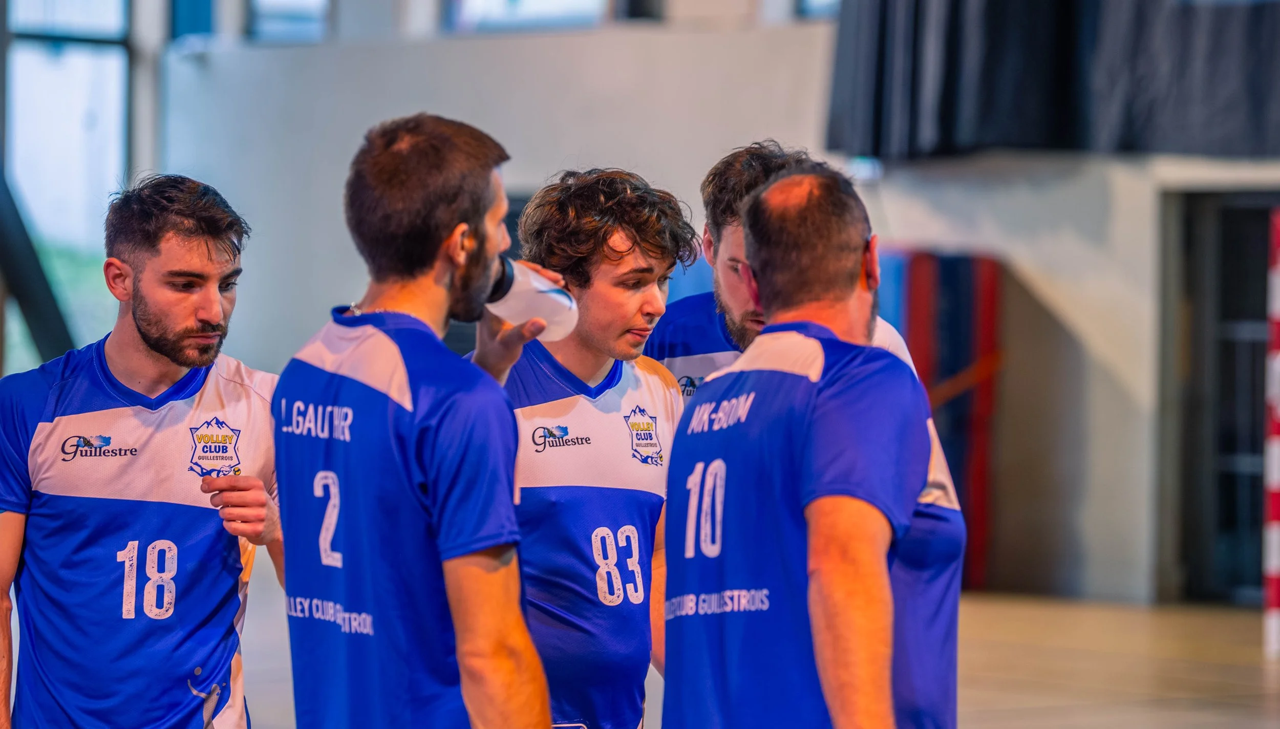 Une équipe de volleyball masculine en uniforme bleu et blanc, apparemment en temps de pause ou en réflexion dans un gymnase.