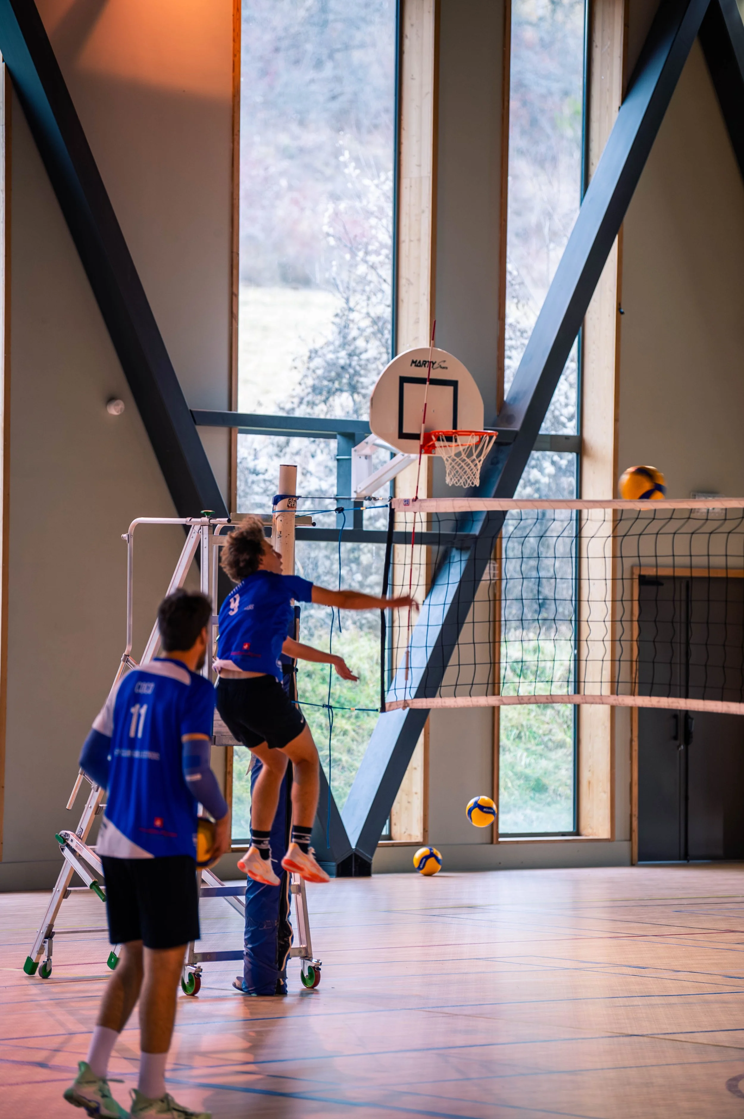 Des joueurs de volley-ball sont en train de jouer dans un gymnase intérieur, avec deux de eux en train de sauter pour atteindre le ballon suspendu au-dessus du filet.