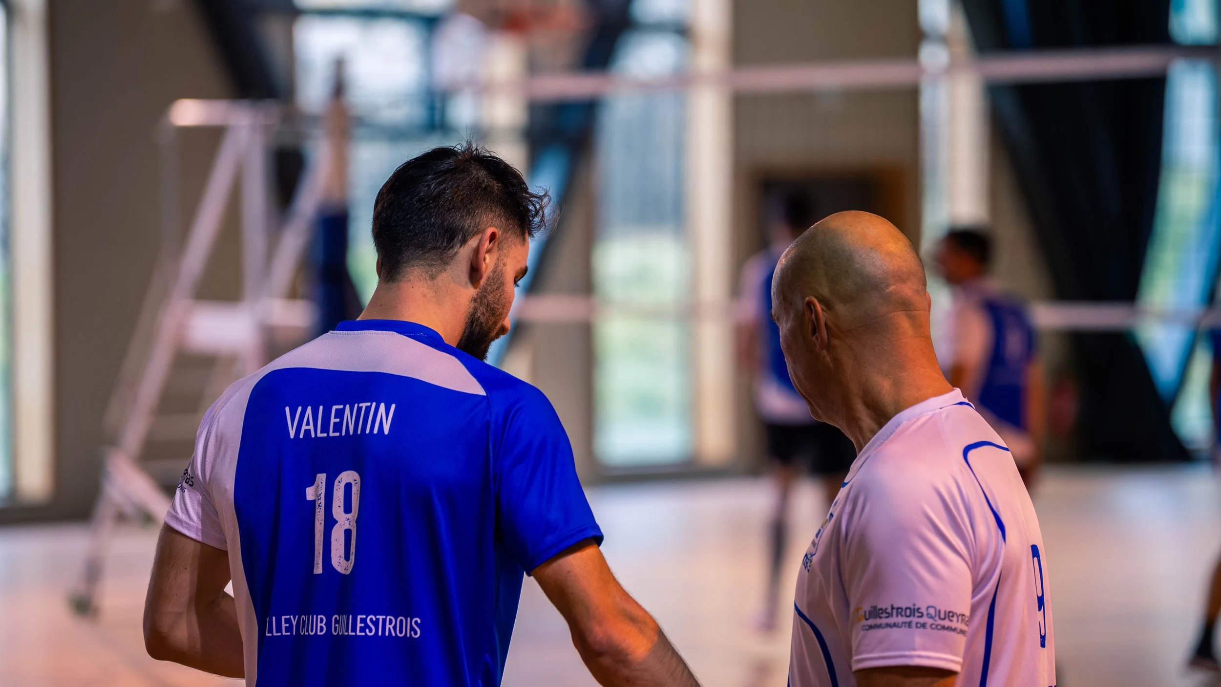 Deux joueurs de volley-ball en tenue de sport discutent dans une salle d'entraînement, avec d'autres personnes en arrière-plan.