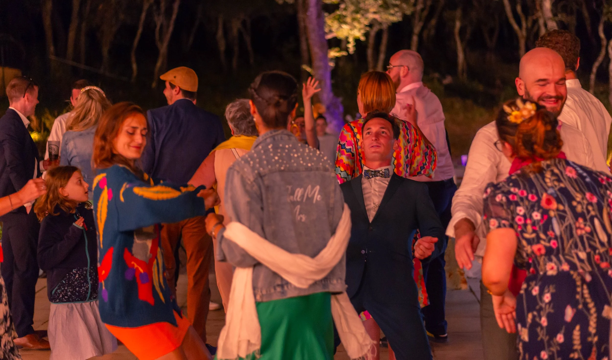 Des personnes dansent et discutent lors d'une fête en plein air la nuit, avec des arbres en arrière-plan et des lumières colorées.