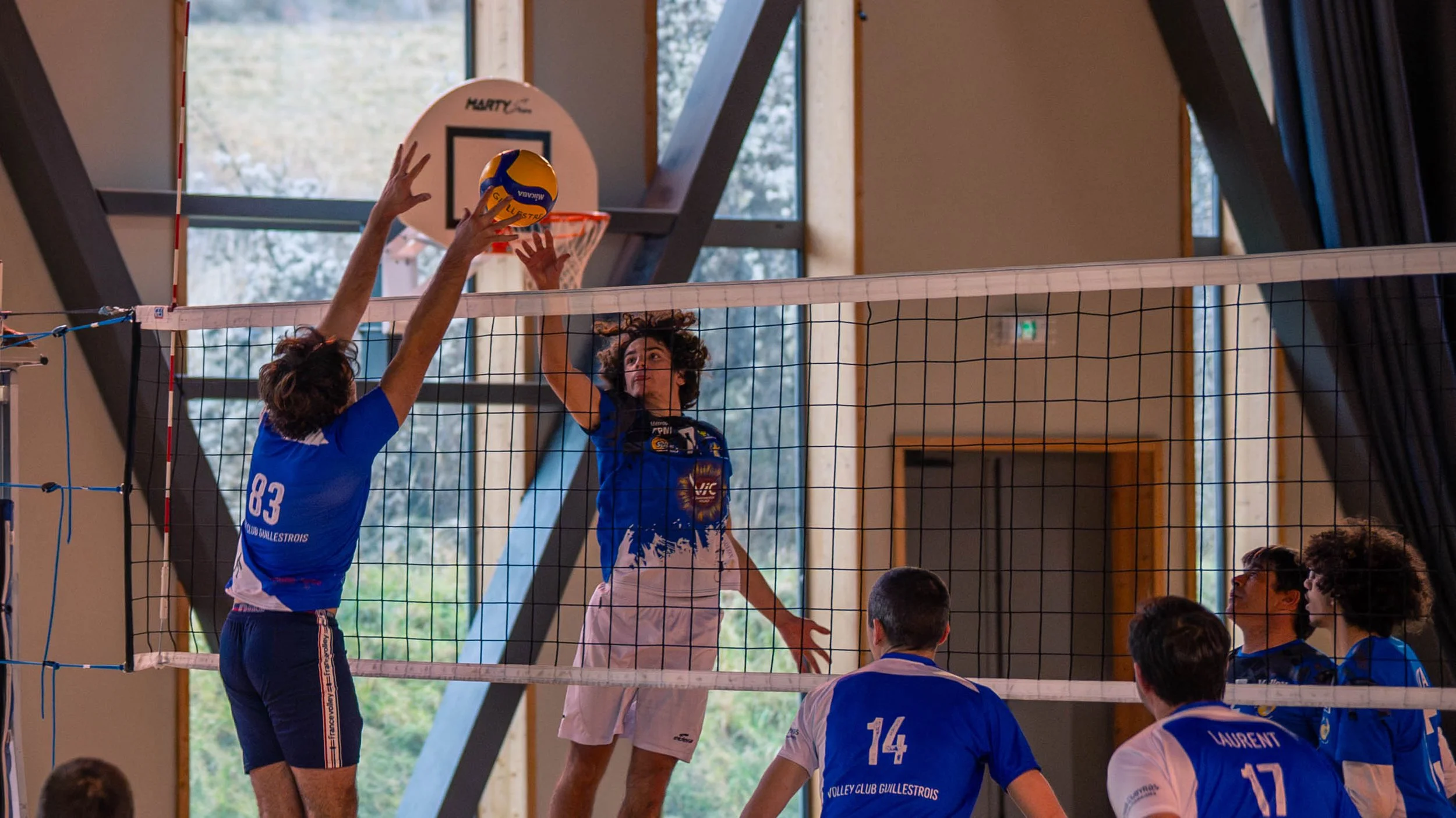 Joueur de volley-ball en action sautant pour attaquer la balle au-dessus du filet, tandis qu'un autre joueur tente de bloquer, dans un gymnase avec fenêtres lumineuses.