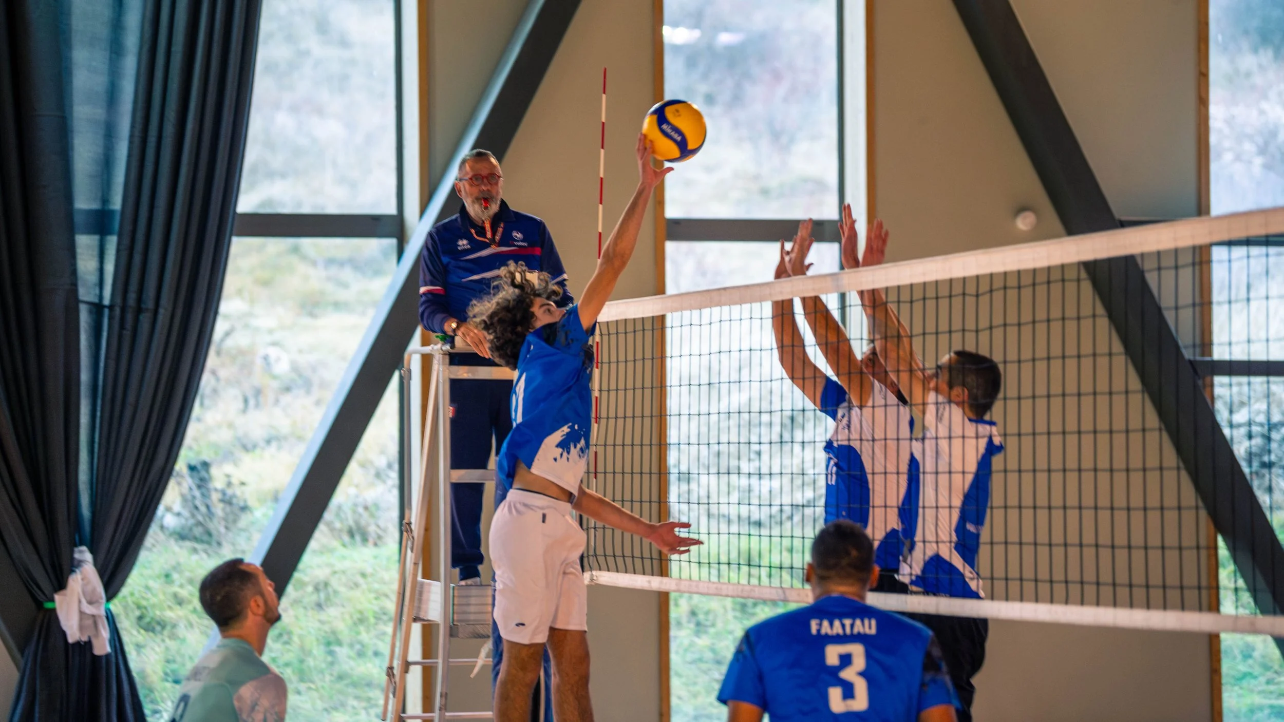 Match de volley-ball en intérieur, un joueur en bleu saute pour frapper le ballon, deux joueurs adverses tentent de bloquer, un arbitre observe depuis une plateforme, spectateurs à l'arrière.