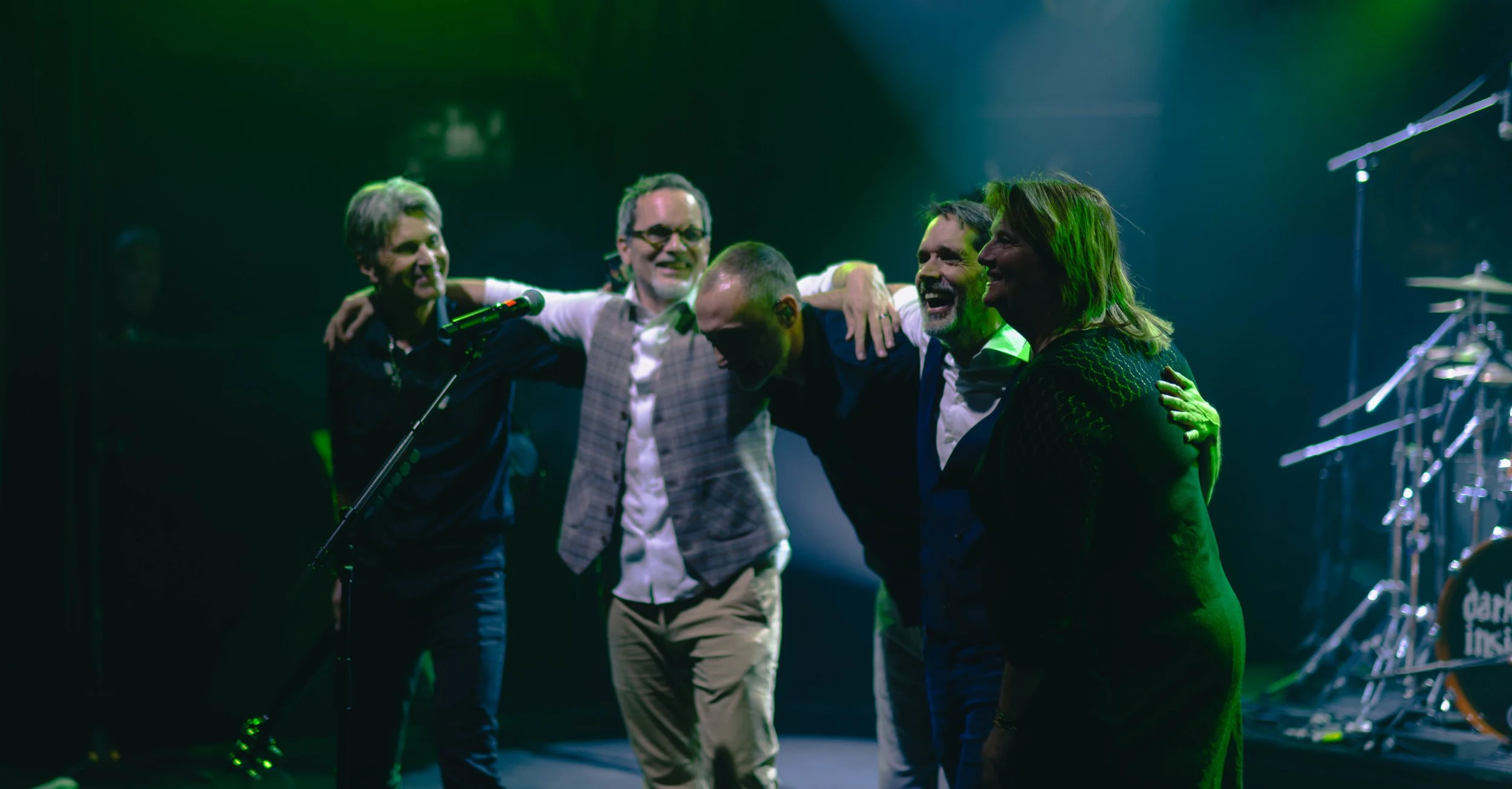 Groupe de cinq personnes souriantes et joyeuses en train de célébrer sur scène, avec un microphone et un instrument de musique en arrière-plan.