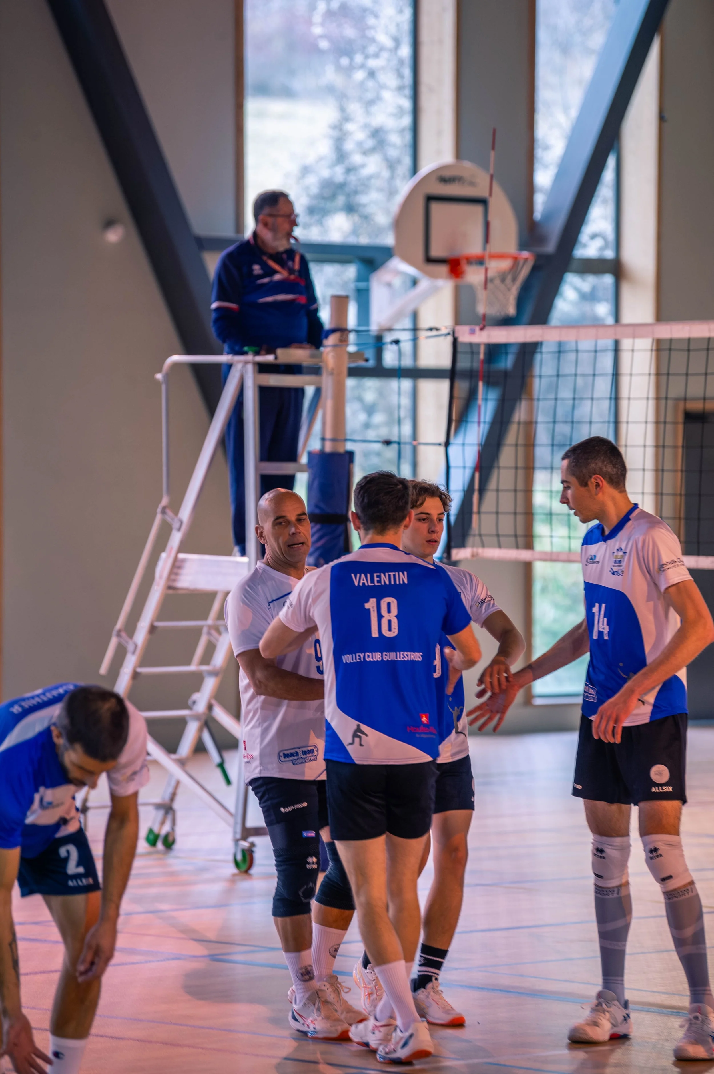 Joueurs de volley-ball en discussion lors d'un match ou entraînement en salle, avec un arbitre ou entraîneur en arrière-plan près du panneau de basket et du filet.