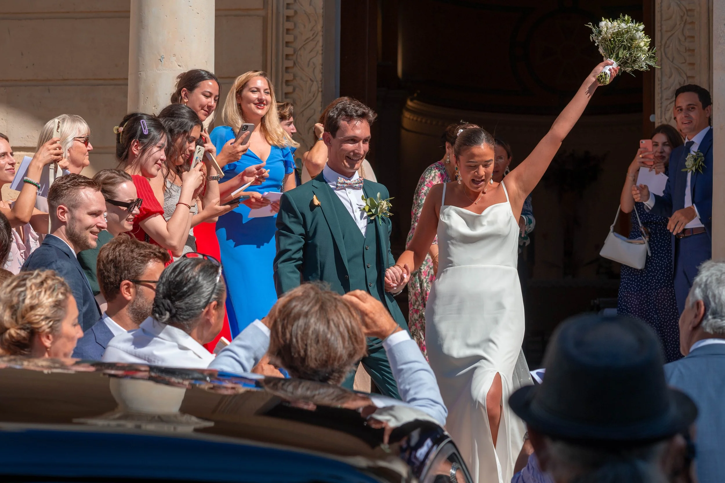 Un couple de mariés marchant main dans la main, entourés d'invités lors d'une cérémonie de mariage en plein air, la mariée brandissant un bouquet en l'air.