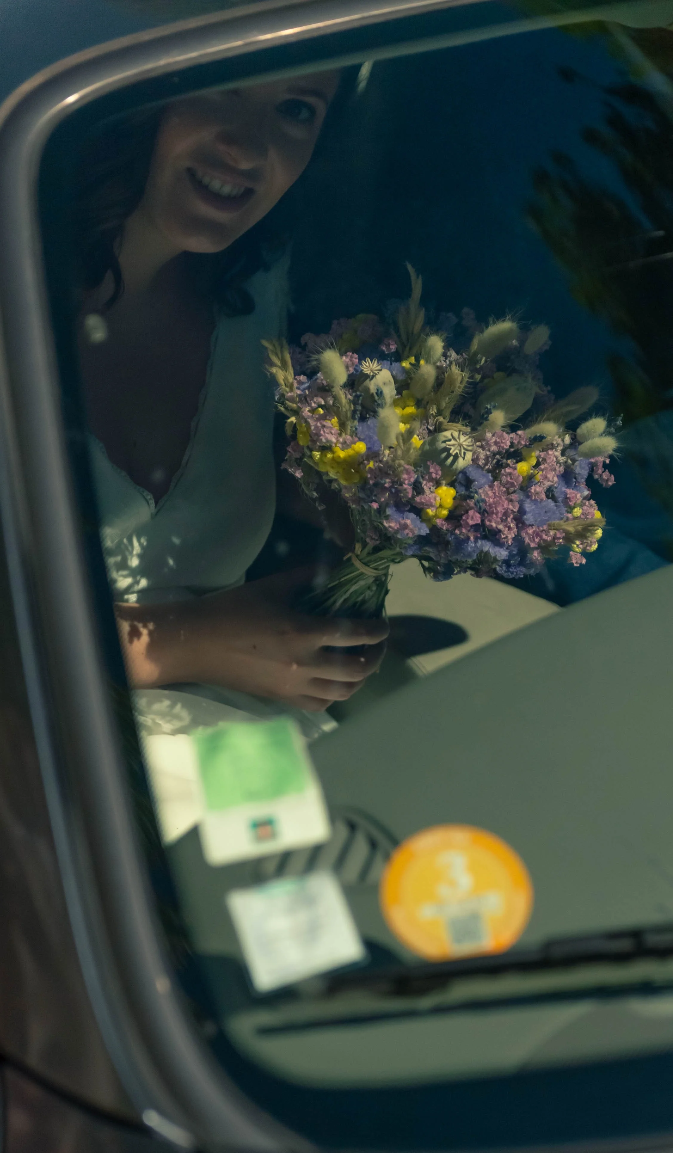 Une femme souriante regarde à travers un miroir de voiture, tenant un bouquet de fleurs multicolores.