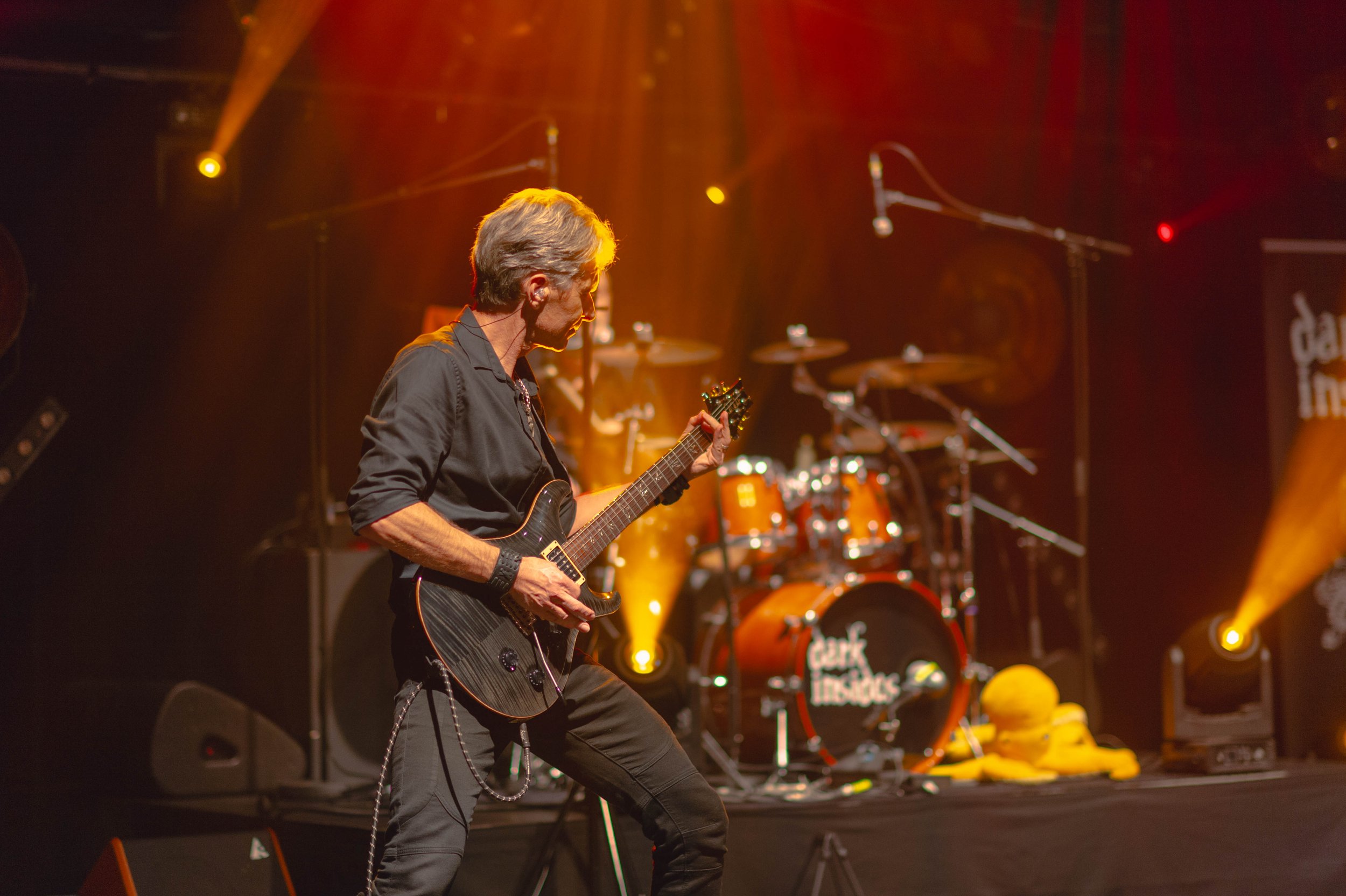 Musicien jouant de la guitare électrique sur scène avec batterie en fond, éclairage chaud.
