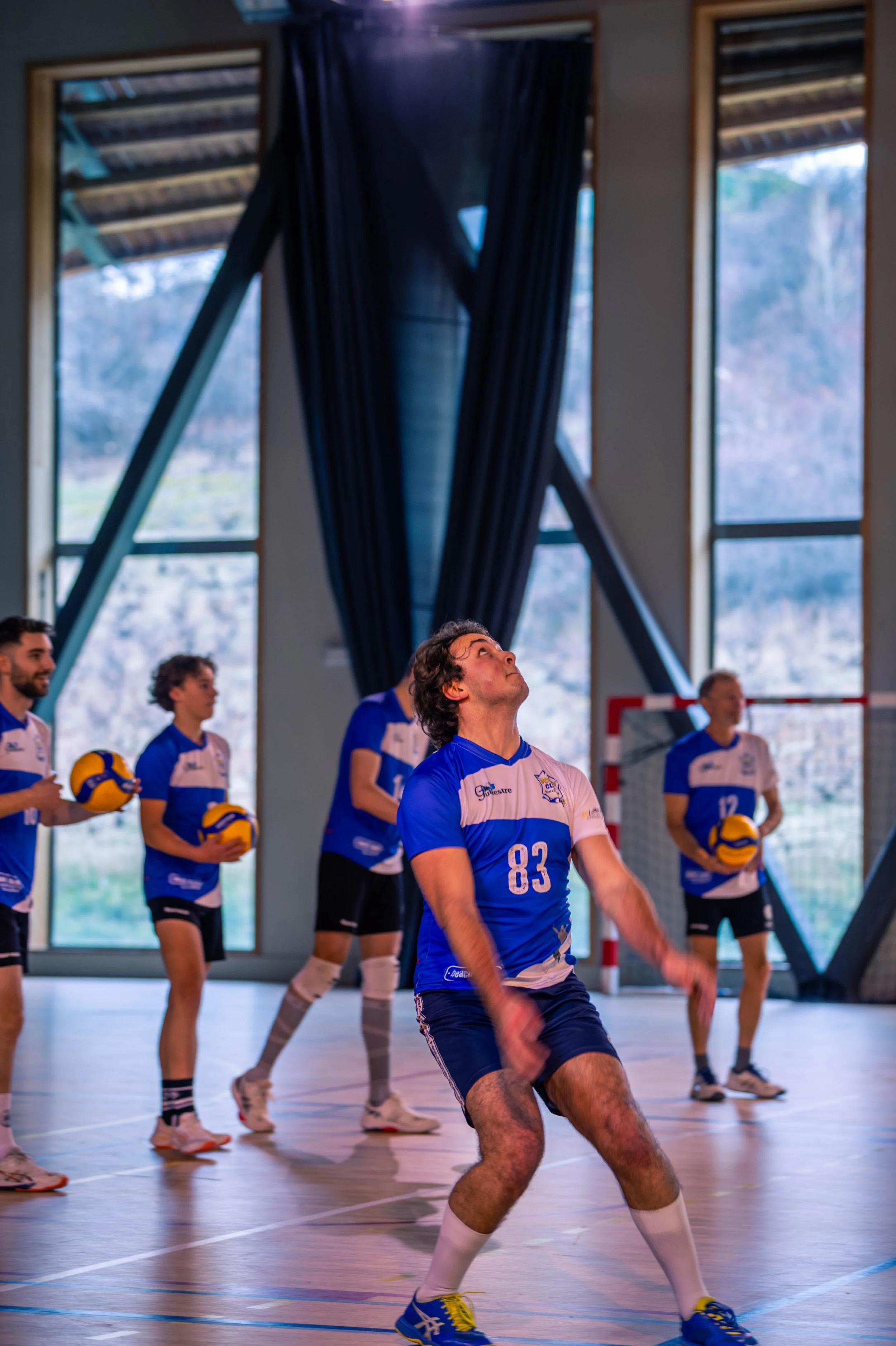 Une équipe de volleyball en train de s'entraîner dans un gymnase avec de grandes fenêtres, portaient des maillots bleus et blancs et tiennent des ballons de volley. La scène est animée, avec un joueur au centre en position prête, prêt à servir ou à r