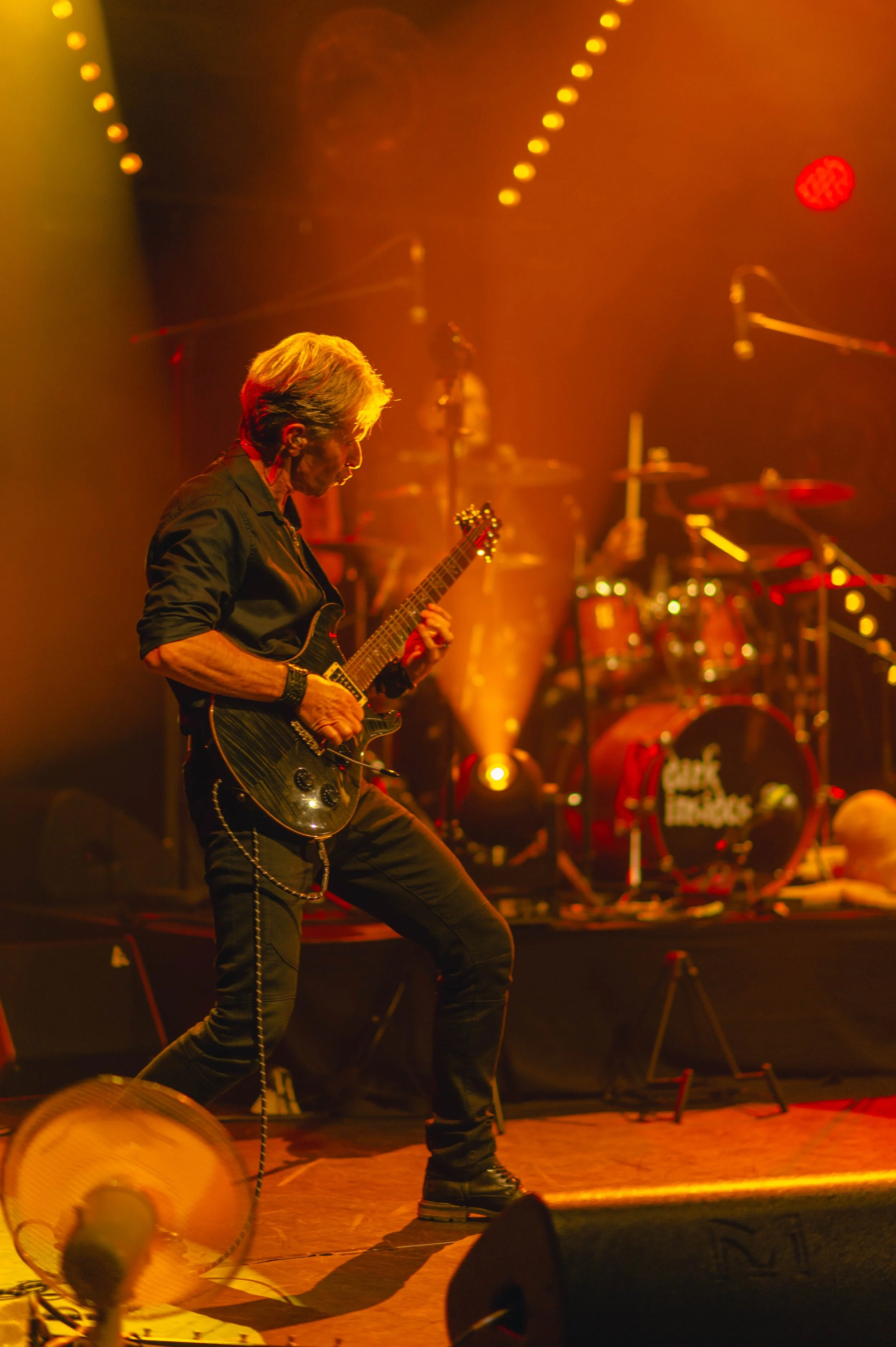 Un guitariste joue sur scène avec un fond d'éclairage orange et rouge, accompagné d'une batterie au arrière.
