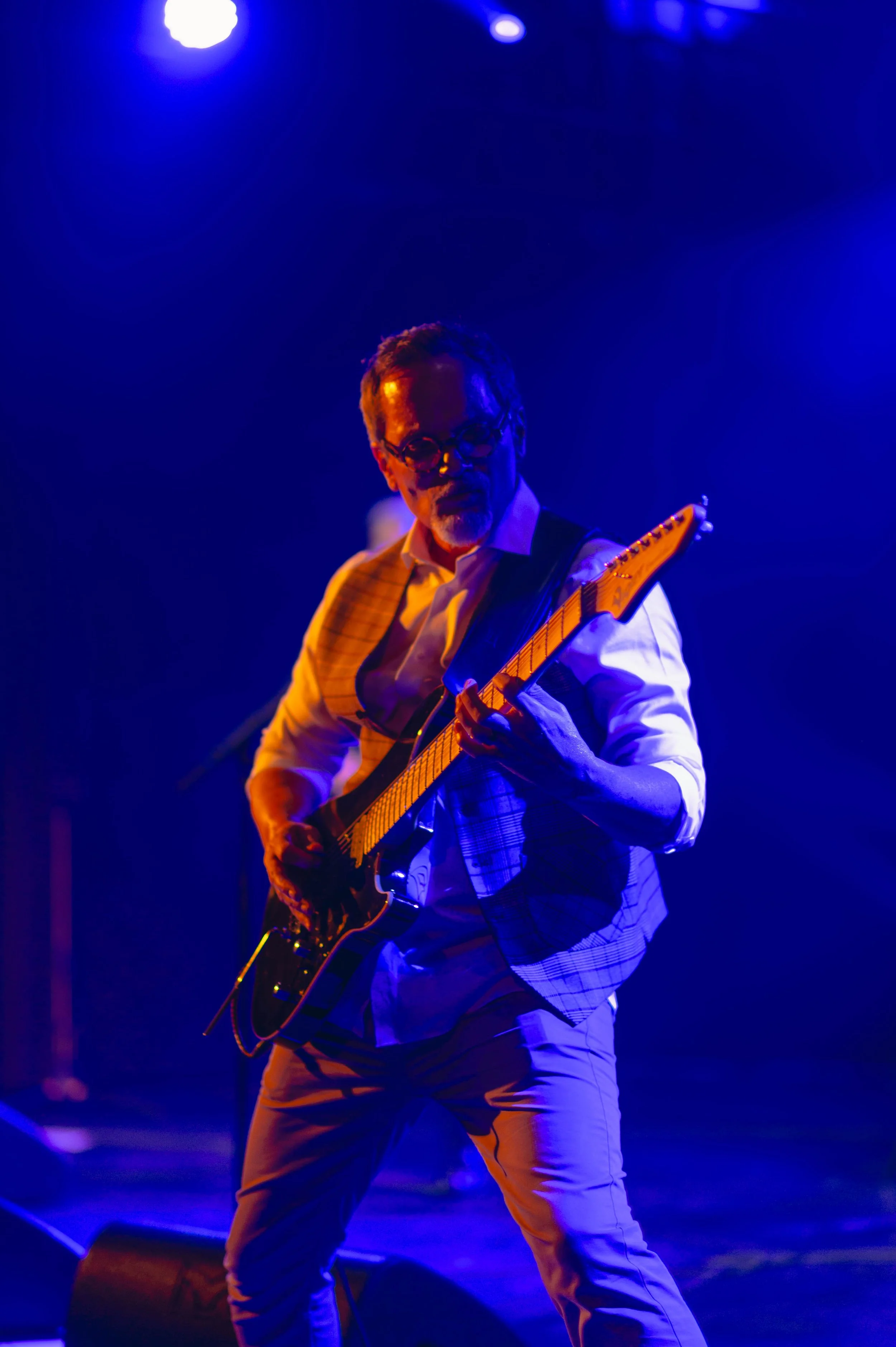 Un homme joue de la guitare électrique sur scène, éclairé par des lumières bleues, portant une chemise blanche et un veston à rayures.